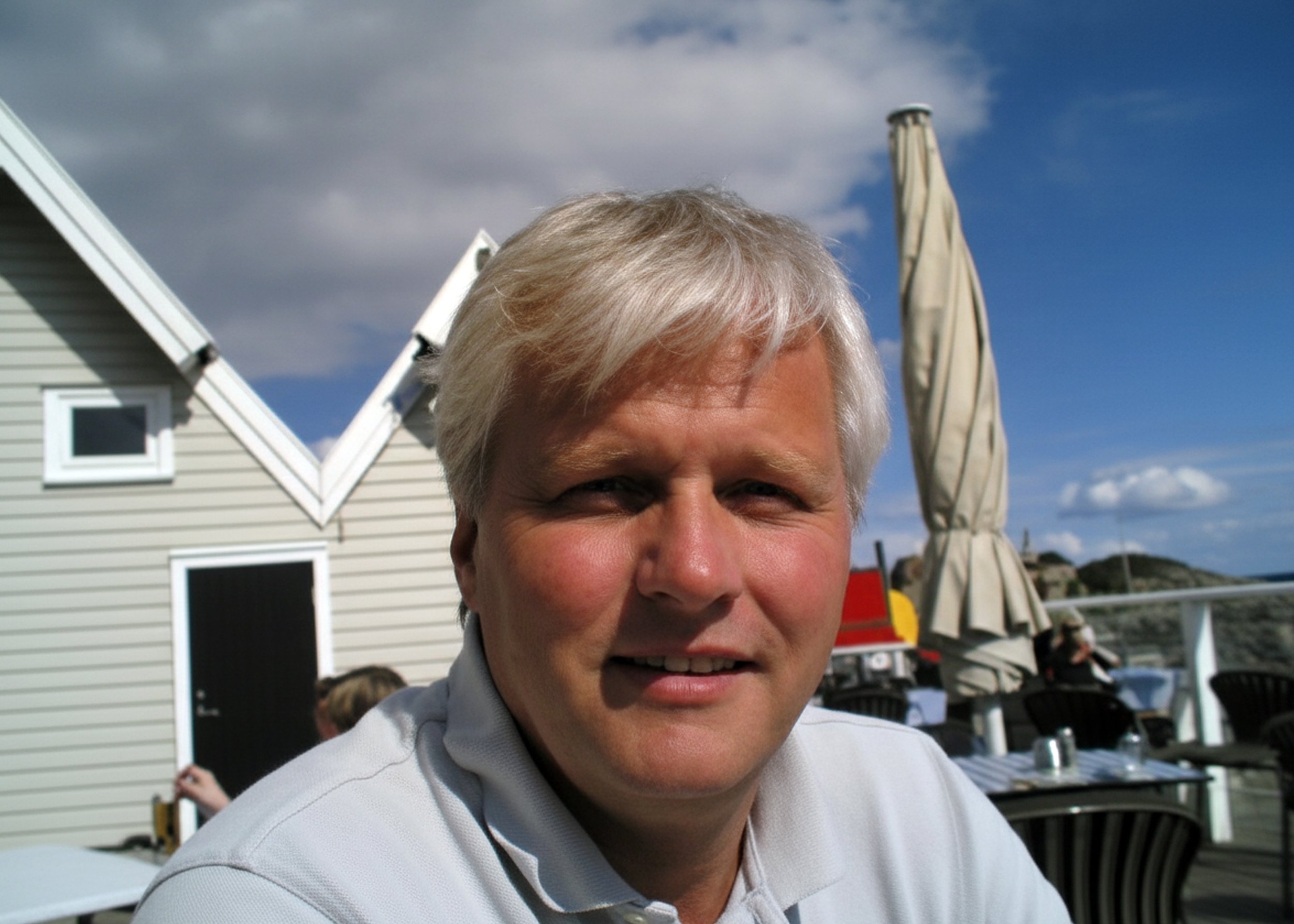 A man with gray hair and a light-colored shirt sitting outdoors at a restaurant or cafe on a sunny day with a building and umbrellas in the background.