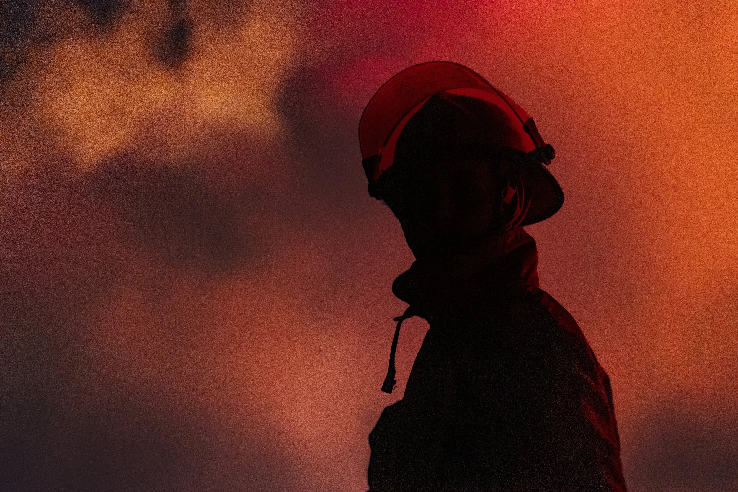 Silhouette of a firefighter wearing a helmet against a smoky, reddish sky.