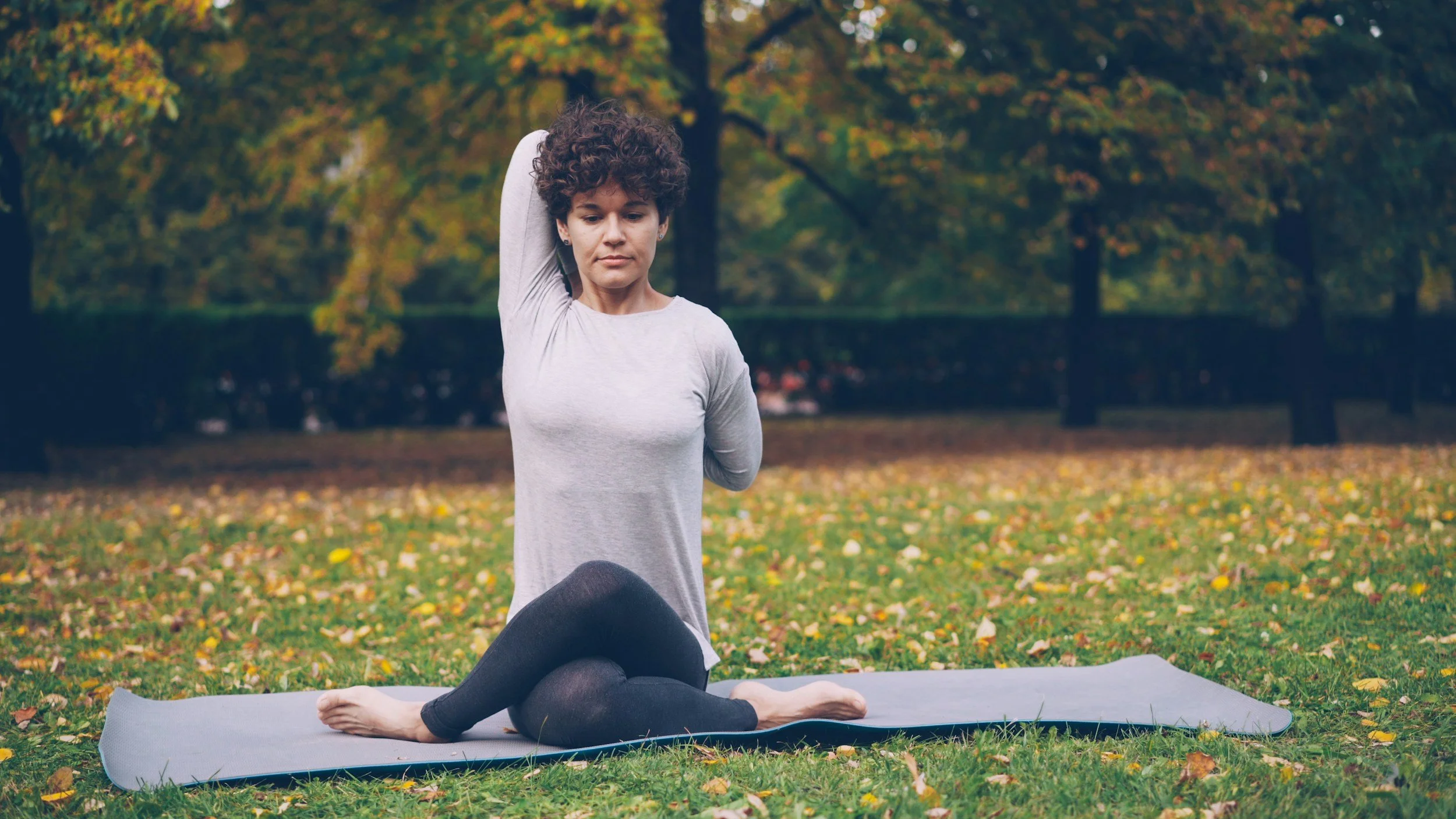 Eine Frau beim Yoga im Park, sie sitzt auf einer Yogamatte im Schneidersitz, umgeben von Bäumen mit herbstlichem Laub.