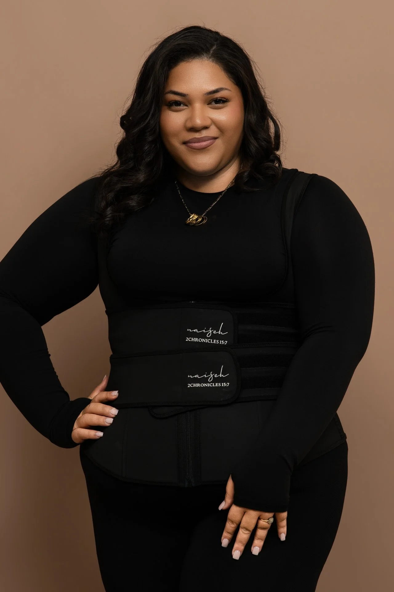 A woman wearing a black long-sleeve top, with natural makeup and dark wavy hair, standing against a plain beige background. She has a gold necklace and is wearing a black waist trainer with the brand name 'naiseh' and scripture '2 Chronicles 15:7' printed on it.