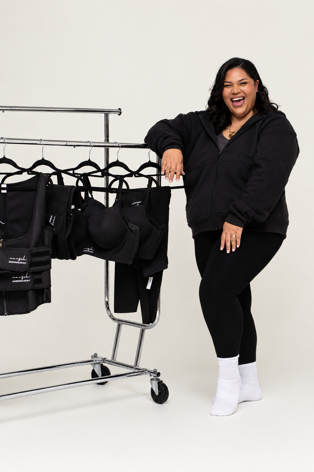 A happy woman in a black hoodie and black leggings standing next to a clothing rack filled with black athletic wear and accessories.