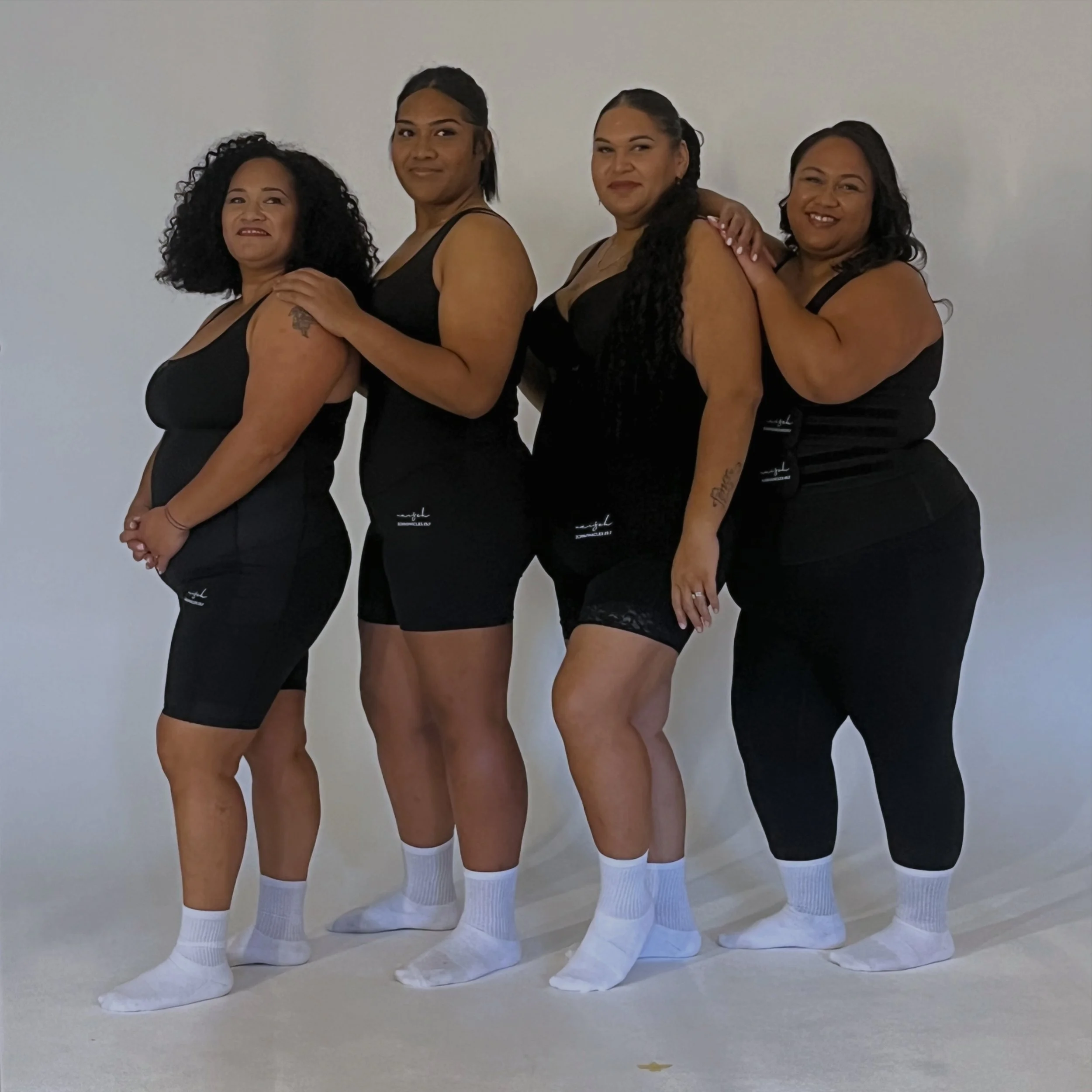 Four women standing in a row against a plain neutral background, wearing black tank tops, black shorts, white knee-high socks, and white athletic shoes, looking at the camera.