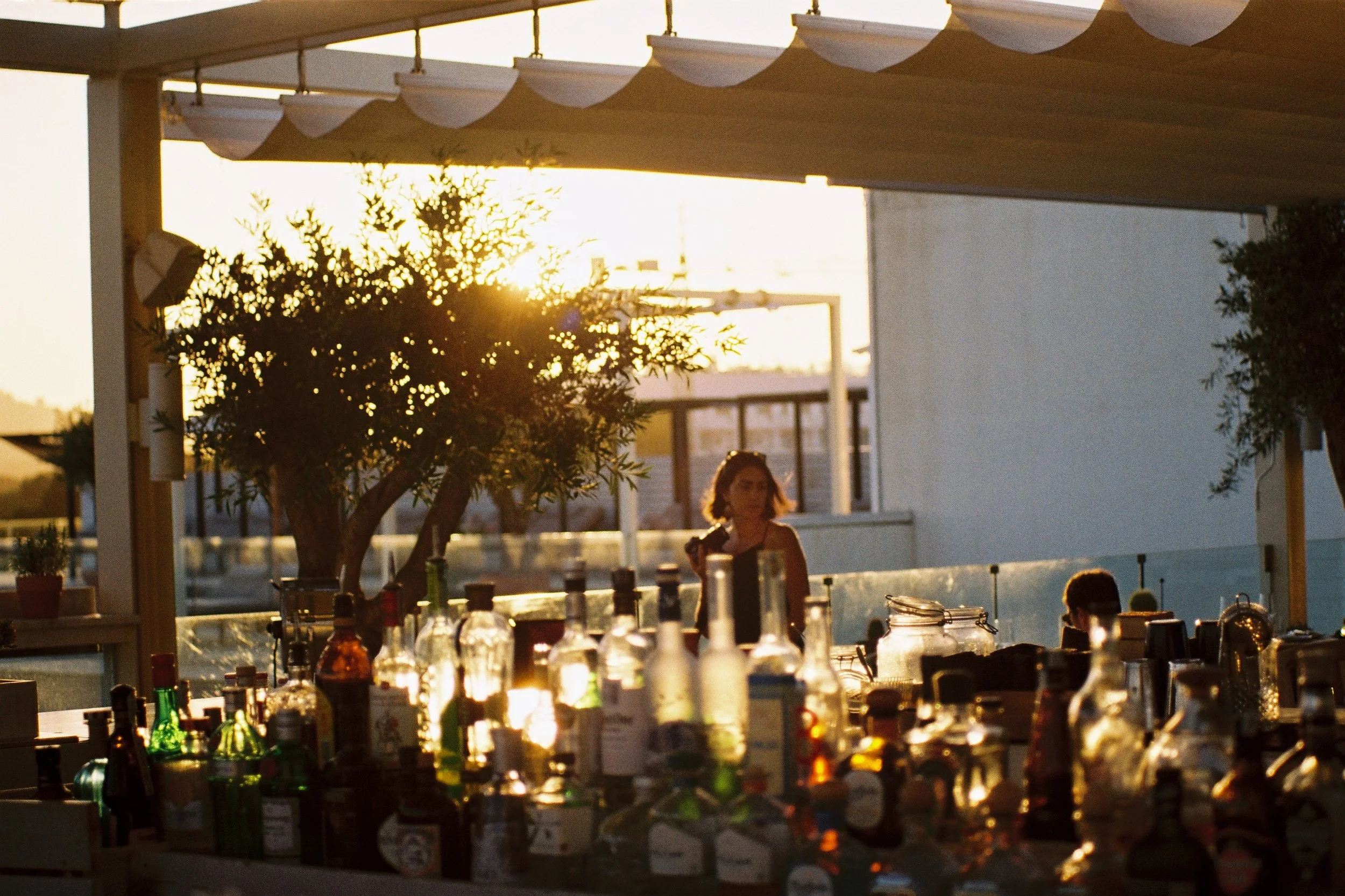 Bar with bottles and barware outdoors during sunset, with trees and structures in the background.