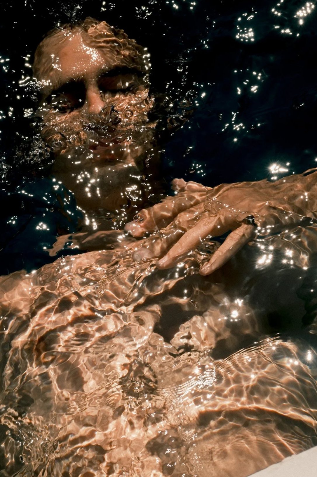 A person with blonde hair floating face down in a dark pool of water, with sunlight reflecting on the surface.