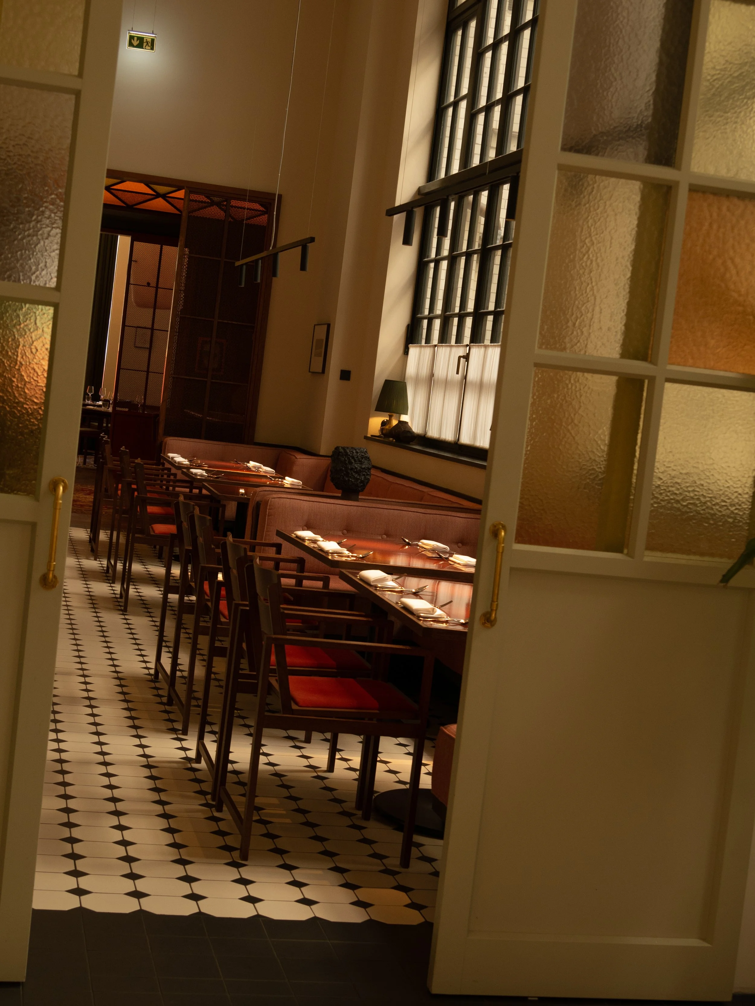 A view inside a restaurant seen through a partially open door with textured glass panels. The interior features a long dining table set with plates and cutlery, a patterned tile floor, large windows with black frames letting in natural light, a green table lamp, and a dark decorative object on a ledge. There is an exit sign above, and dark wood accents and partitioning inside.