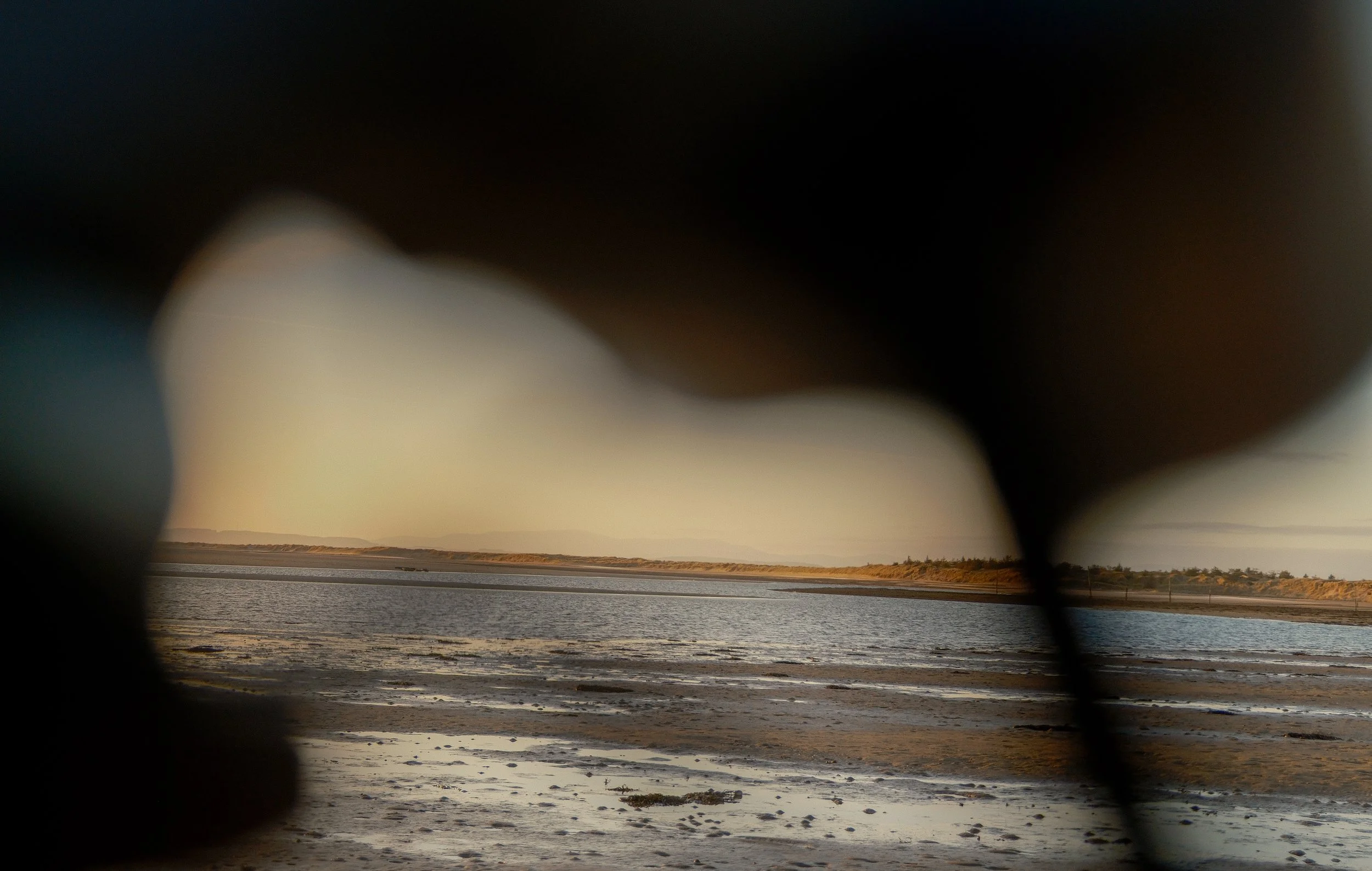 A view of a body of water and distant shoreline through a dark, blurry frame that appears to be a hole or opening.