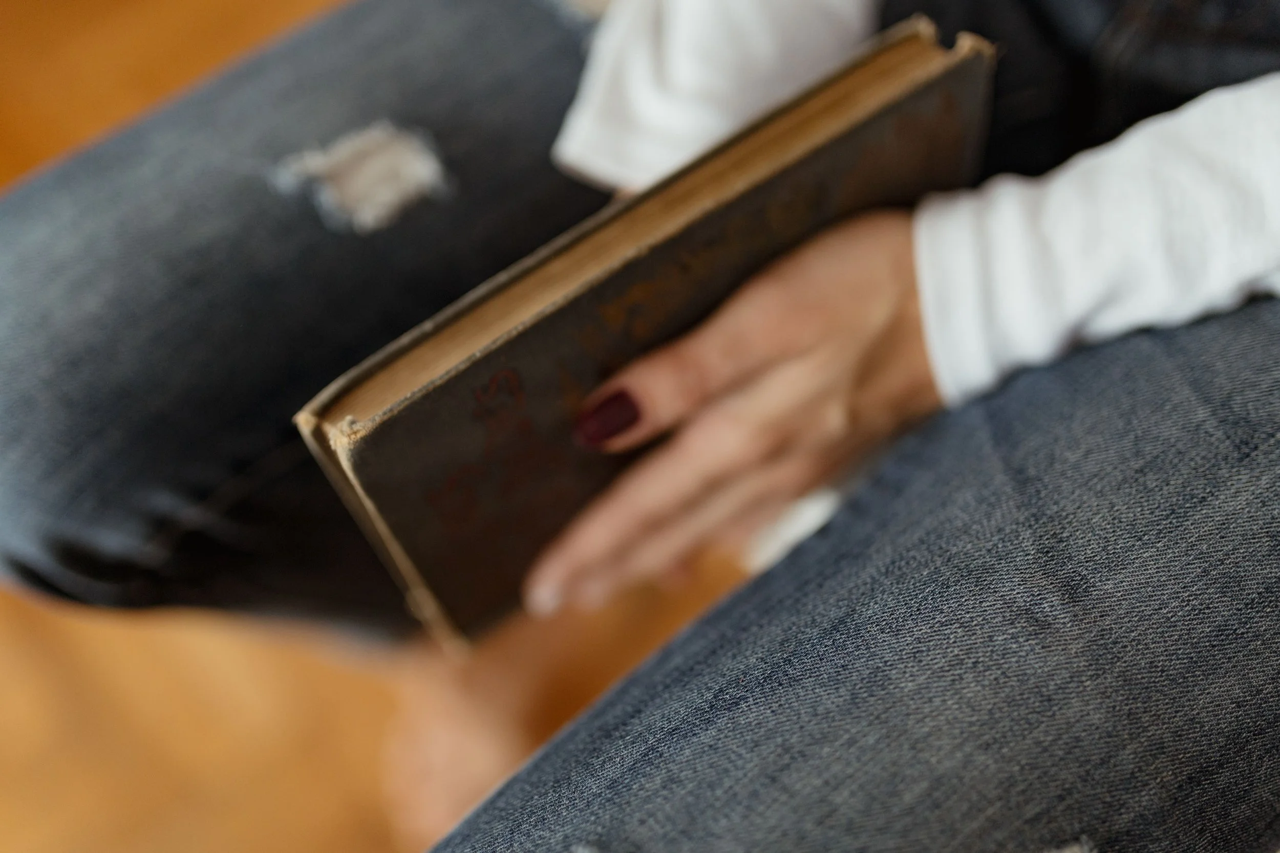A person sitting on a surface with dark jeans, holding a small antique book with a worn dark cover. The person's hand has dark nail polish.