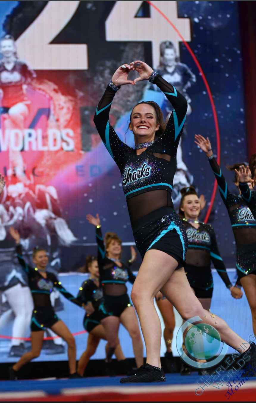 Cheerleaders performing at a competition, cheerleader wearing a black and blue sparkle uniform with the word 'Sharks' on it. The background displays the stage of the Cheerleading Worlds competition. Therapist Mackenzie