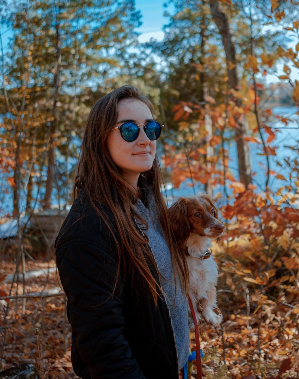 A woman with long brown hair wearing round blue sunglasses, a gray hoodie, and black jacket, holding a small brown and white dog outdoors in a wooded area with autumn leaves.