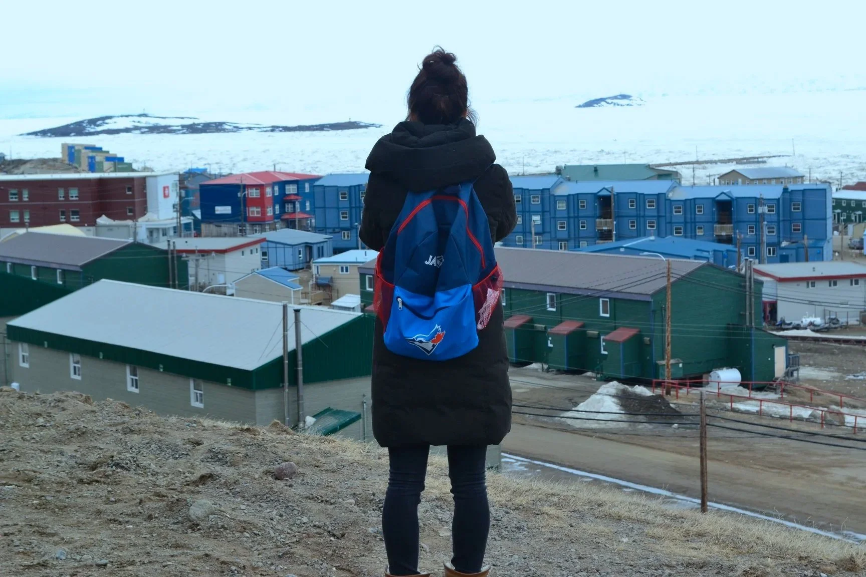 Person standing on a dirt hill overlooking a town with colorful houses, some snow, and mountains in the background.