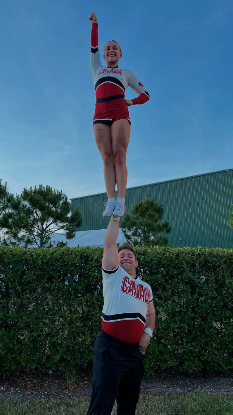 A male cheerleader holding a female cheerleader on his hand while she stands on his palm outdoors with a hedge, trees, and a green building in the background, both wearing Canada-themed uniforms.