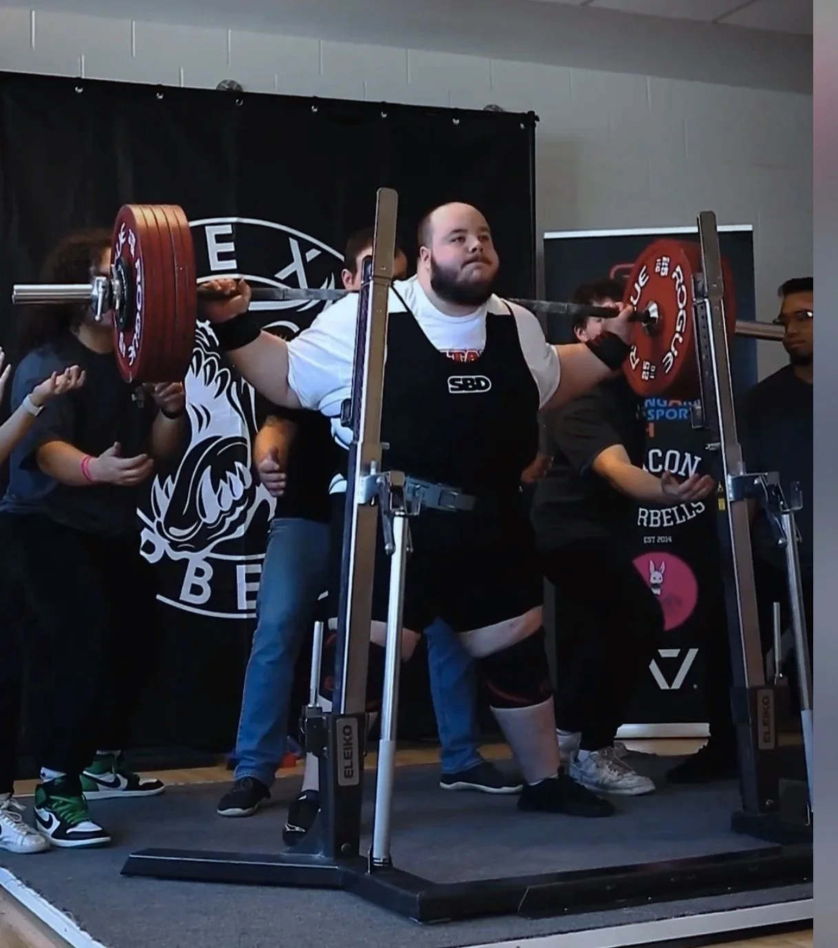 A man performing a squat with a barbell loaded with red weights, surrounded by people at a weightlifting competition.