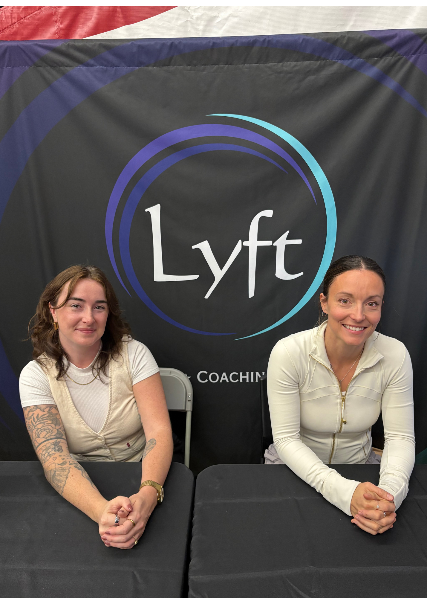 Two women sitting at a table with black tablecloth, smiling, in front of a black backdrop with the Lyft logo and the words 'Lyft Coaching'. Lyft counselling attending a cheerleading event. Information booth.