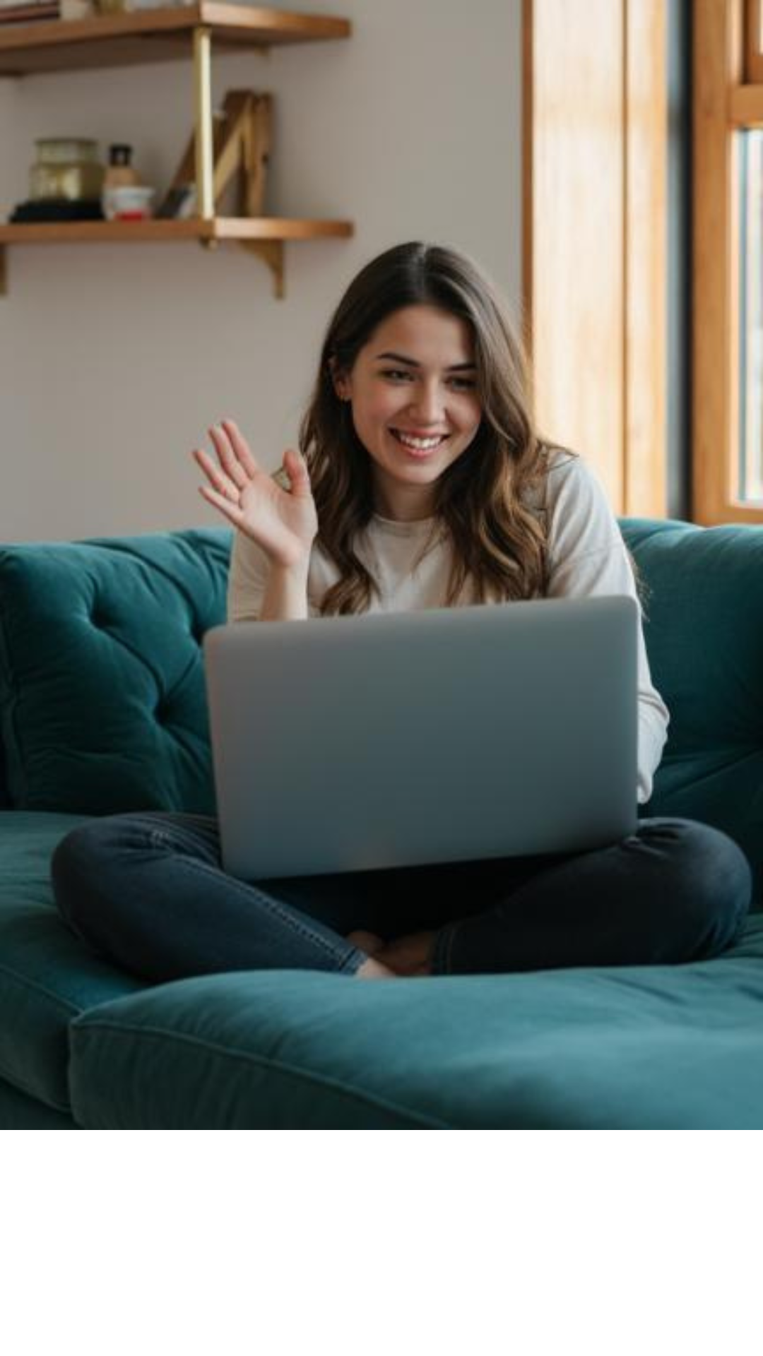 A young woman sitting cross-legged on a teal couch, smiling and waving at her open silver laptop, with a wooden shelf and window in the background.