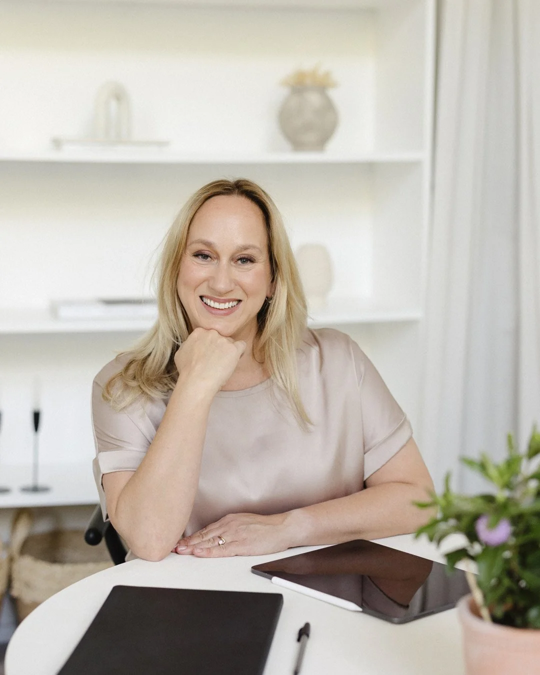 A woman with blonde hair smiling and resting her chin on her hand, sitting at a white table with a tablet, stylus, and potted plant, in a bright room with white shelves and decorative items in the background.