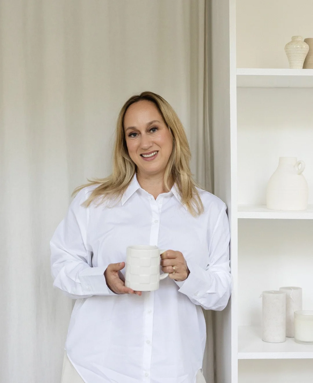 Woman with blonde hair smiling, wearing a white shirt, holding a white ceramic mug, standing in front of a white shelf with white pottery and vases.