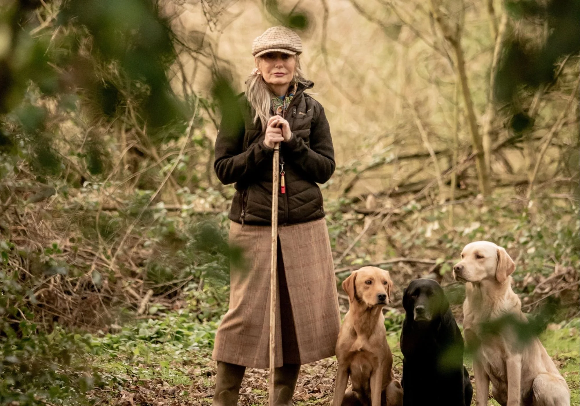 Dawn Scott in outdoor clothing standing in a wooded area holding a walking stick with three dogs sitting beside her.