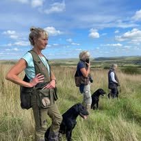 Dawn Scott and clients practicing walked up training with their labrador dogs in a grassy field under a blue sky with clouds.