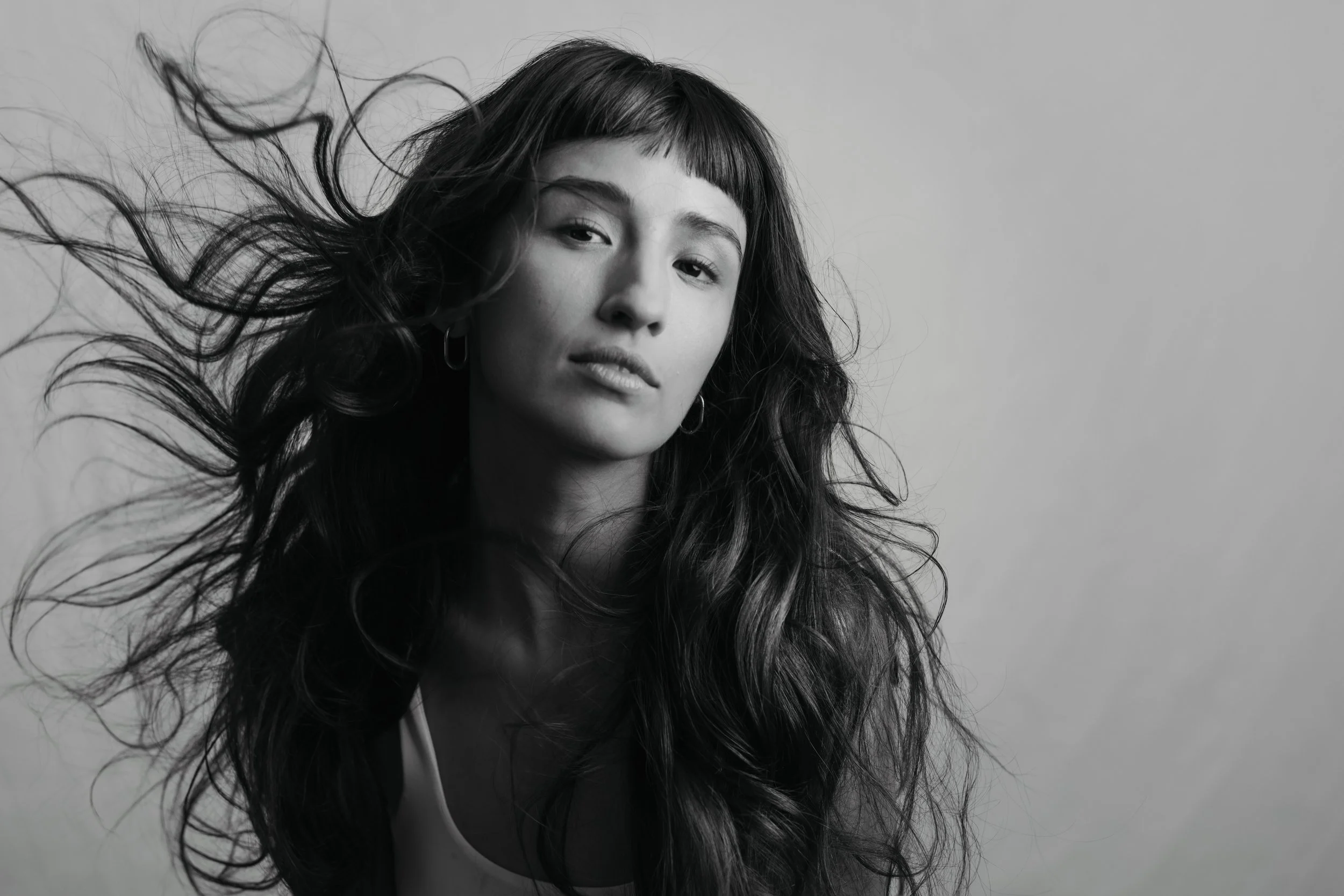 Beauty editorial hair and makeup by Jenna Smith, Berlin.Black and white portrait of a woman with long wavy hair, wearing hoop earrings, looking slightly to the side with a neutral expression, hair blowing to the left against a plain light background.