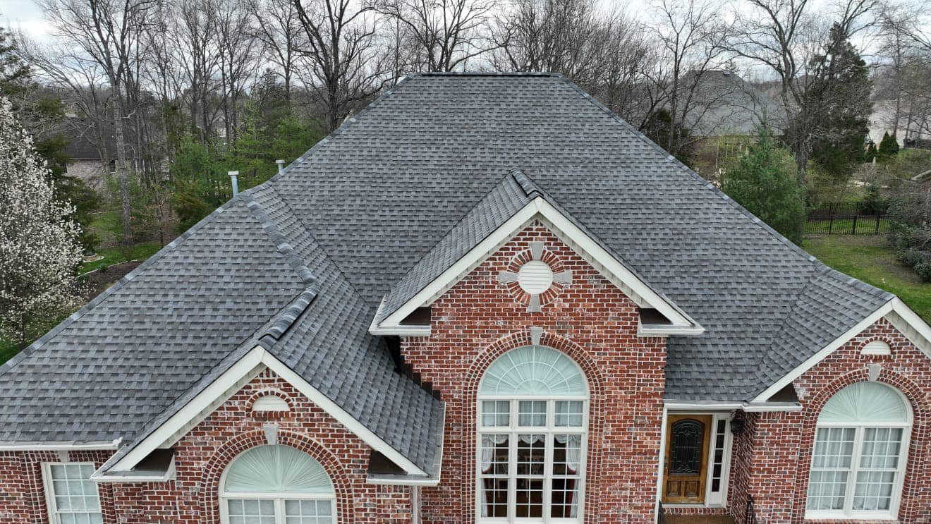 Aerial view of a red brick house with a gray shingled roof, large arched windows, and a front door, surrounded by trees and a backyard.