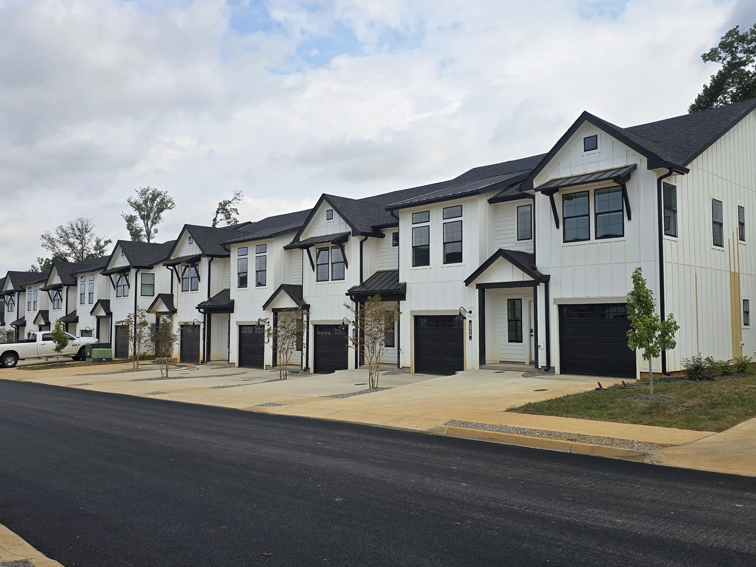 A row of newly built white townhouses with black roofs, large windows, small trees in front, and a black driveway. Overcast sky with some blue sky and clouds.