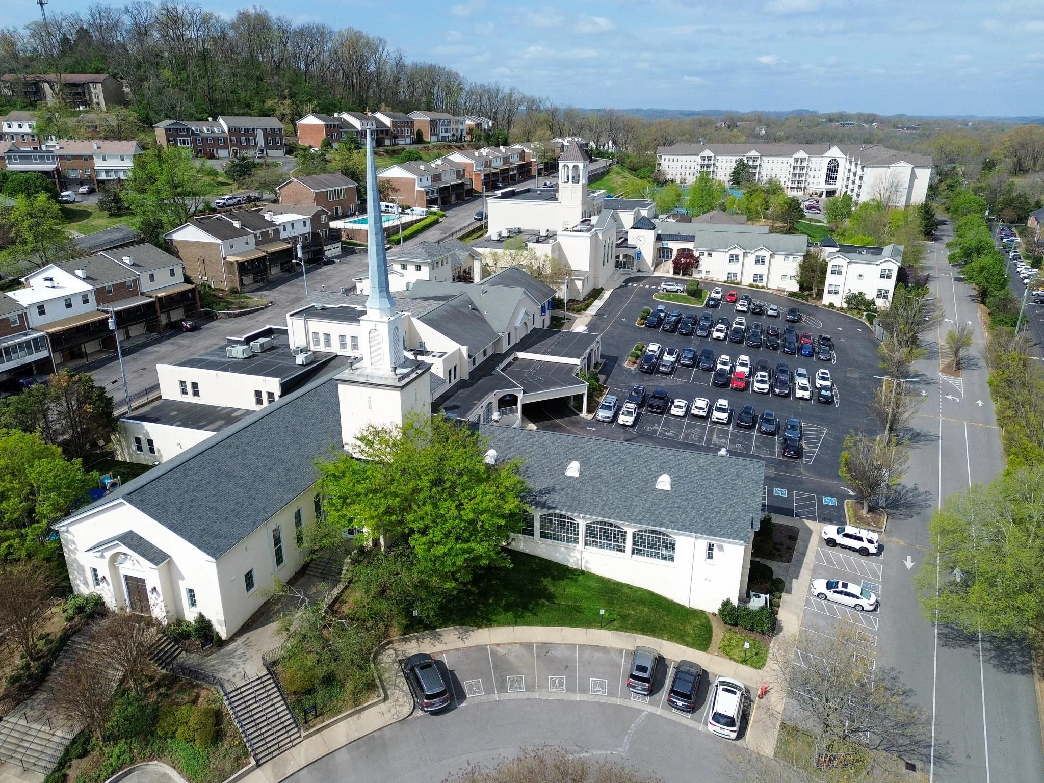 Aerial view of a residential area with multi-story apartments, houses, parking lots, and a church with a tall steeple, surrounded by greenery and trees, on a bright, partly cloudy day.