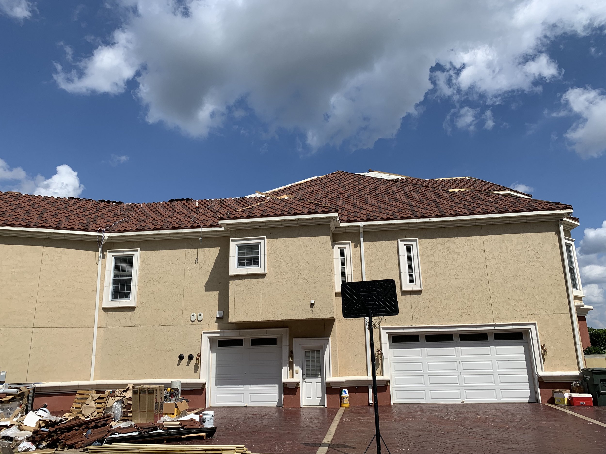 The back of a beige house with a red tiled roof under a blue sky with scattered clouds. There is a basketball hoop in the driveway, some moving boxes, and construction materials on the ground.