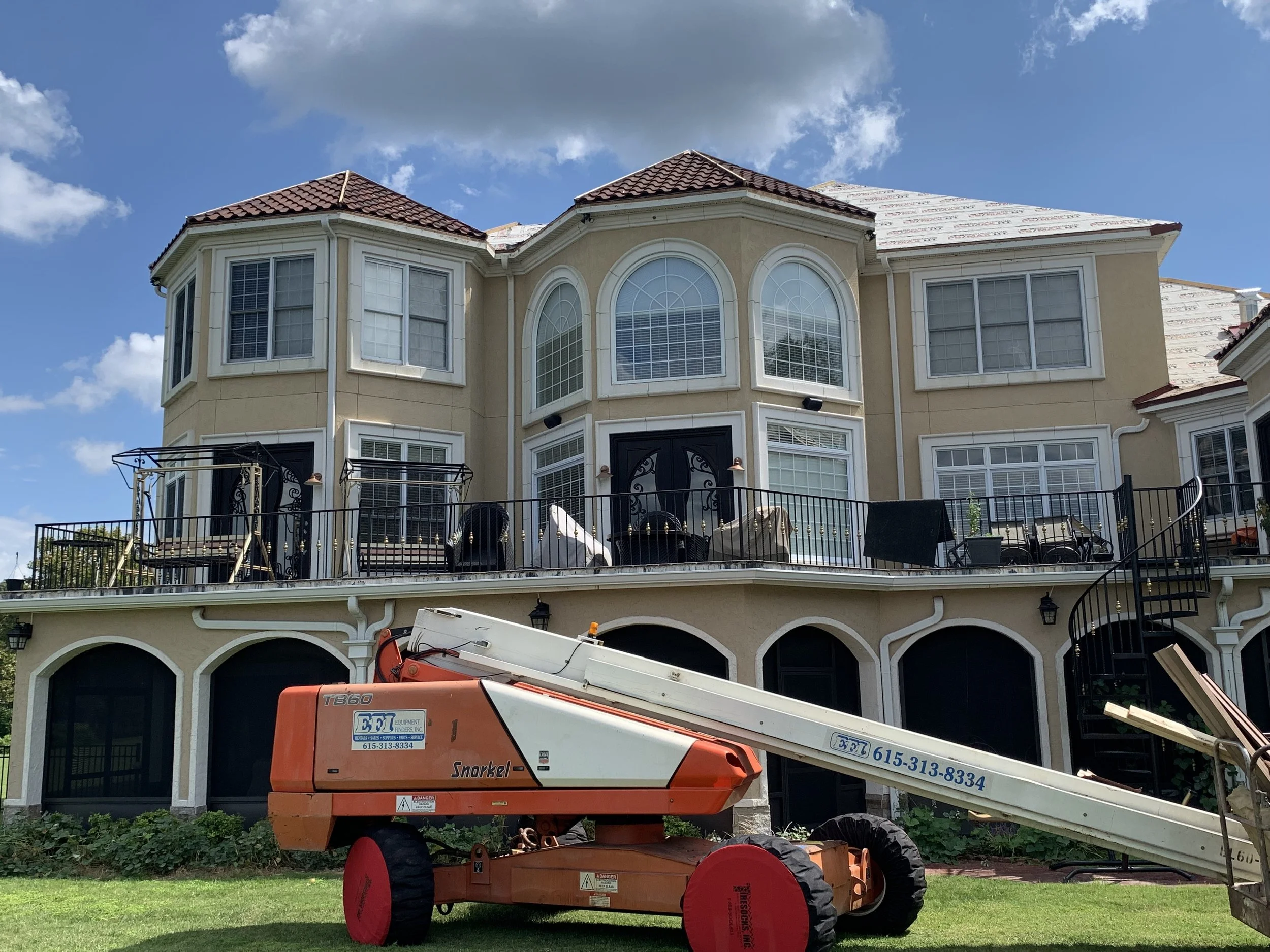 The image shows a large, beige multi-story house with a red tiled roof, multiple arched and rectangular windows, and a balcony with black ornate railings. There is a construction or maintenance lift in front of the house, with the words "EF I Equipme
