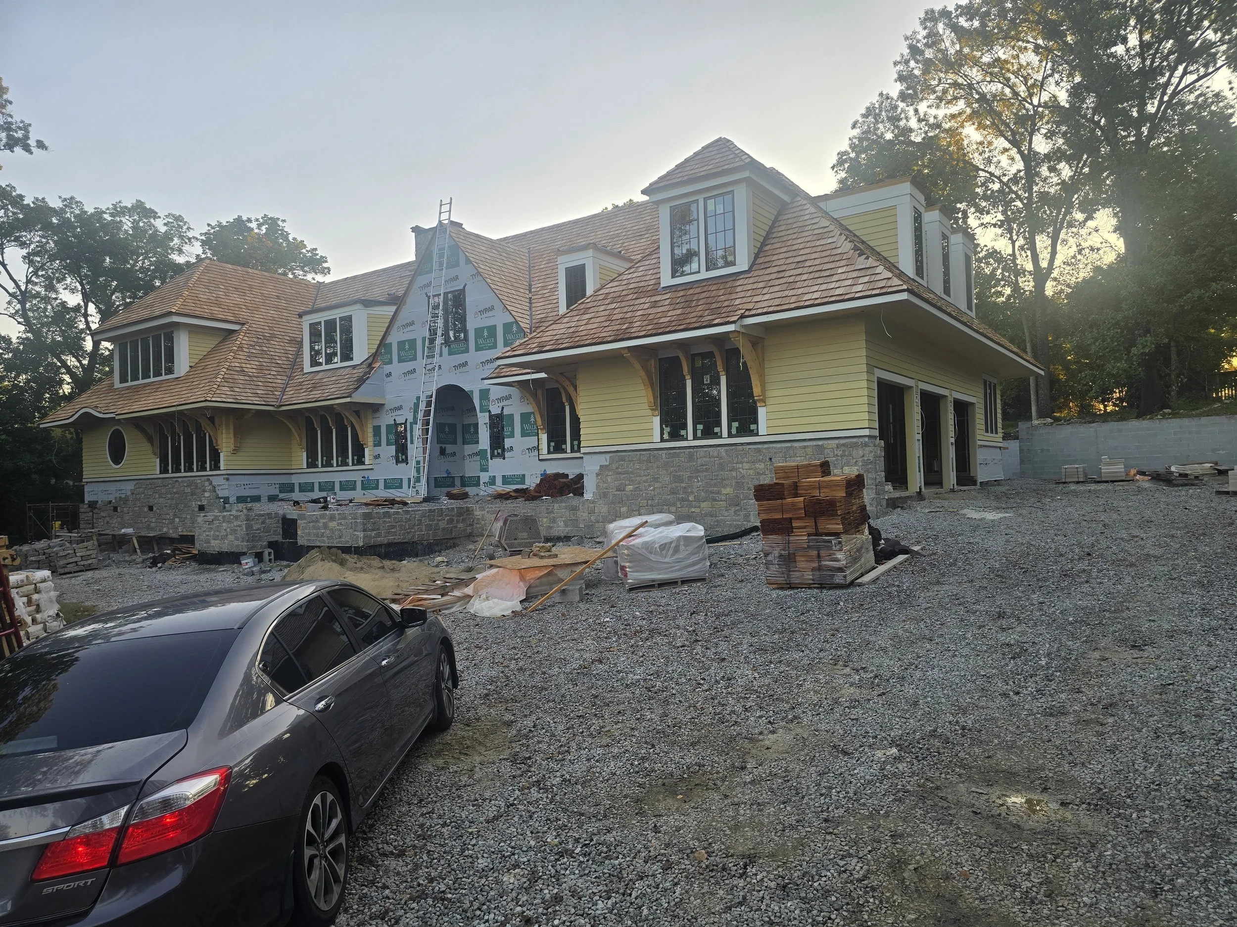 A house under construction with a partially finished exterior, yellow siding, and a stone foundation, surrounded by gravel and construction materials, and a black luxury car parked in the foreground.