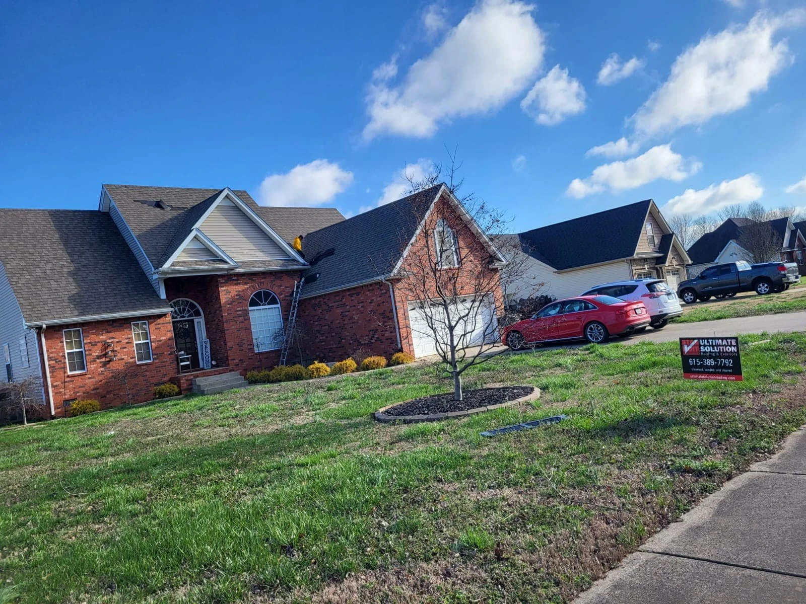 A suburban house with a mix of brick and siding exterior, multiple cars parked in the driveway, a leafless tree in the front yard, and a for sale sign.