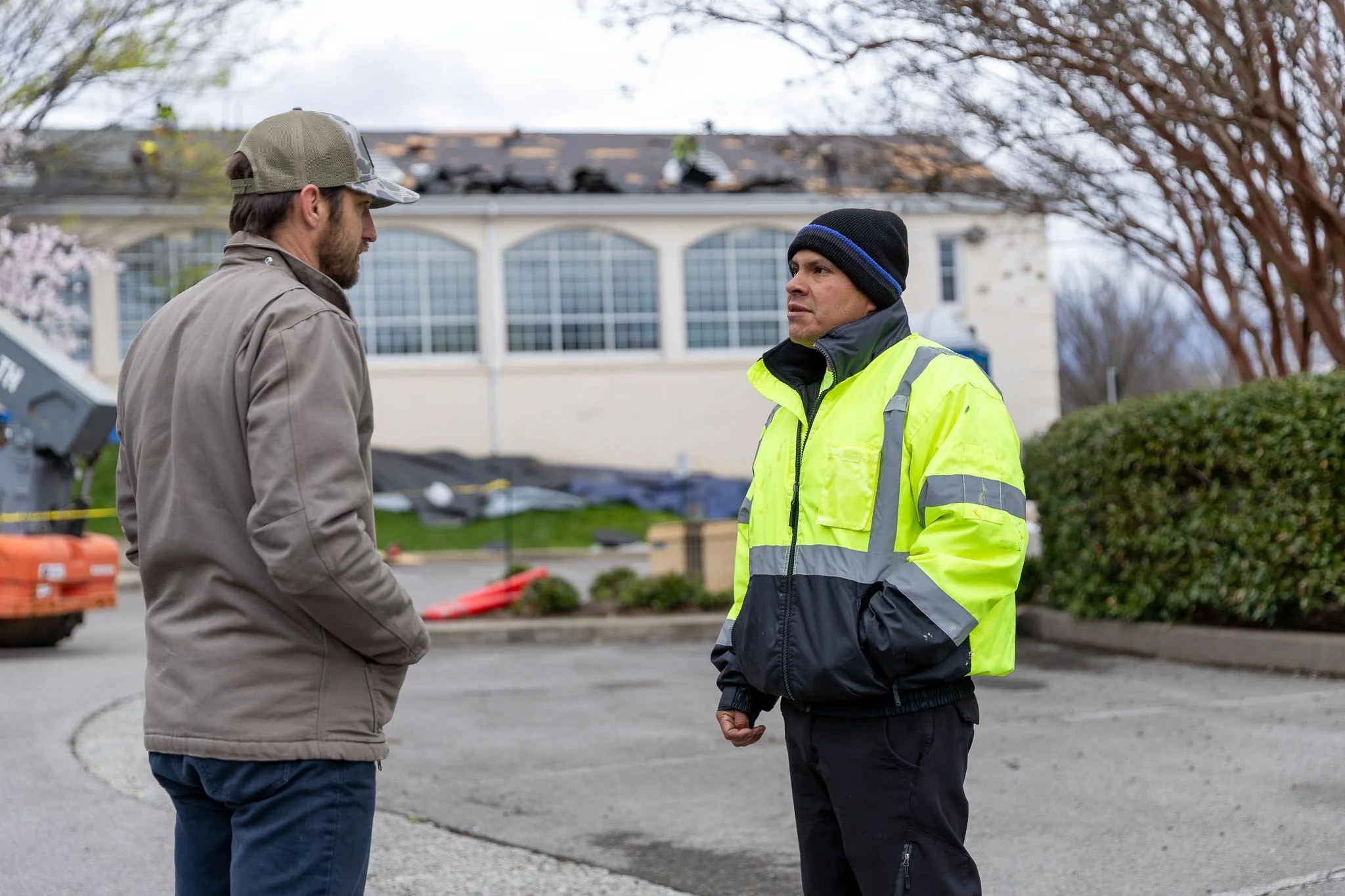 Two men standing outdoors, engaged in conversation. One man wears a camouflage cap and a beige jacket, the other man is in a black beanie and a high-visibility yellow jacket. Background includes a multi-story building with large windows and leafless trees.