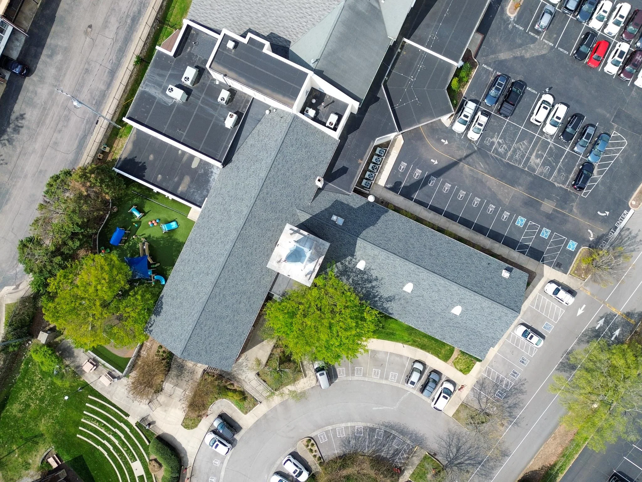 An aerial view of a parking lot adjacent to a building, with cars parked indicates and a designated area for bicycles, as well as surrounding trees and pathways.