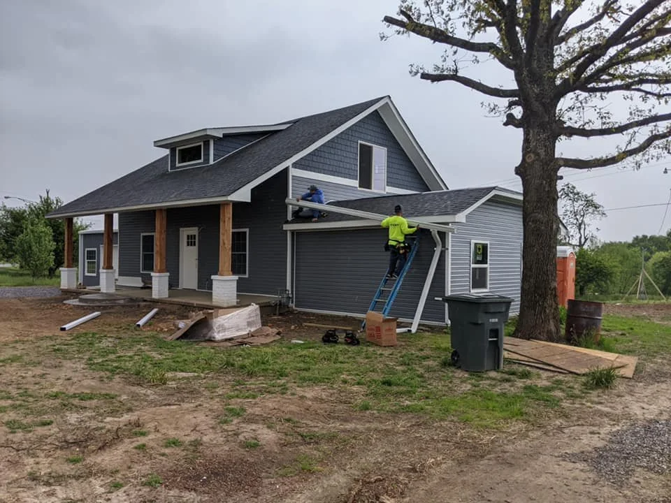 Construction workers installing a new roof on a blue house with a porch, surrounded by dirt and construction materials, near a large tree.