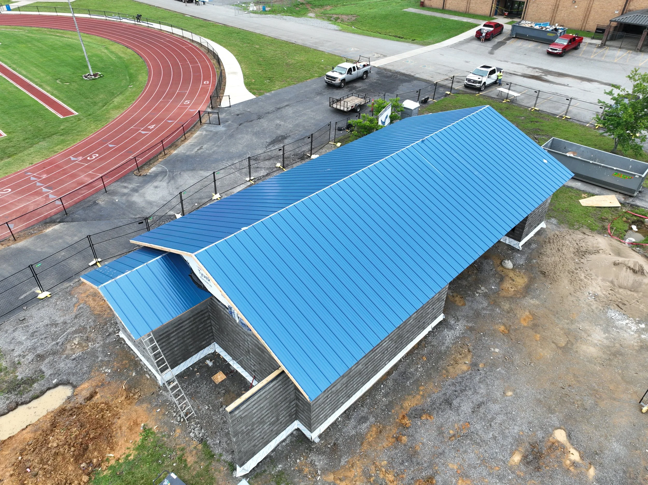 Construction site near a running track with a blue-roofed structure and multiple parked vehicles in the background.