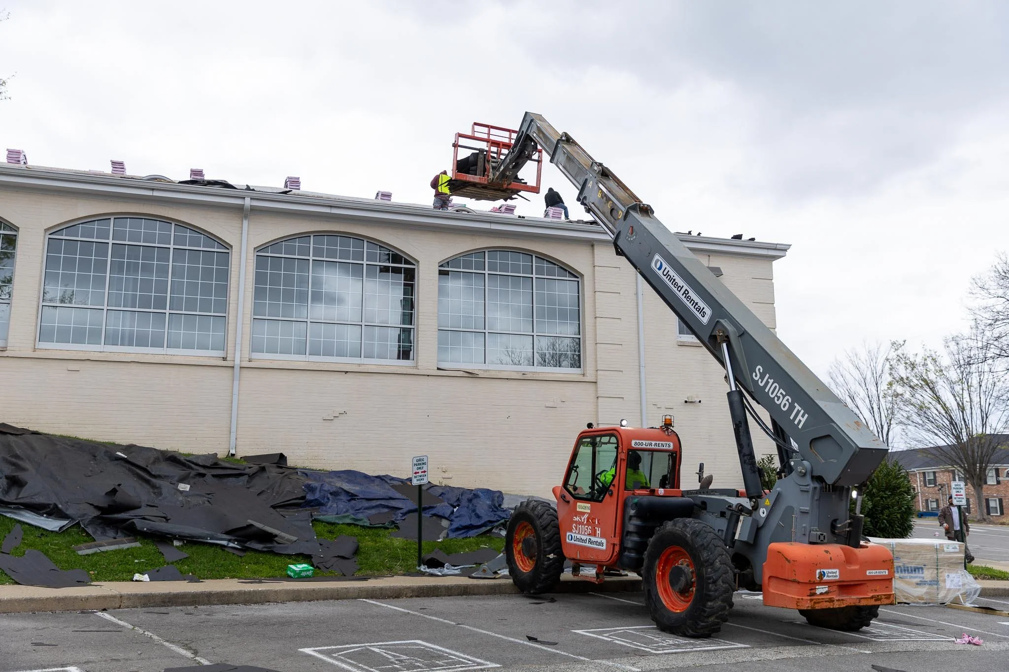 Construction workers on the roof of a building working with roofing materials, using a lift and a crane to position materials on the roof.