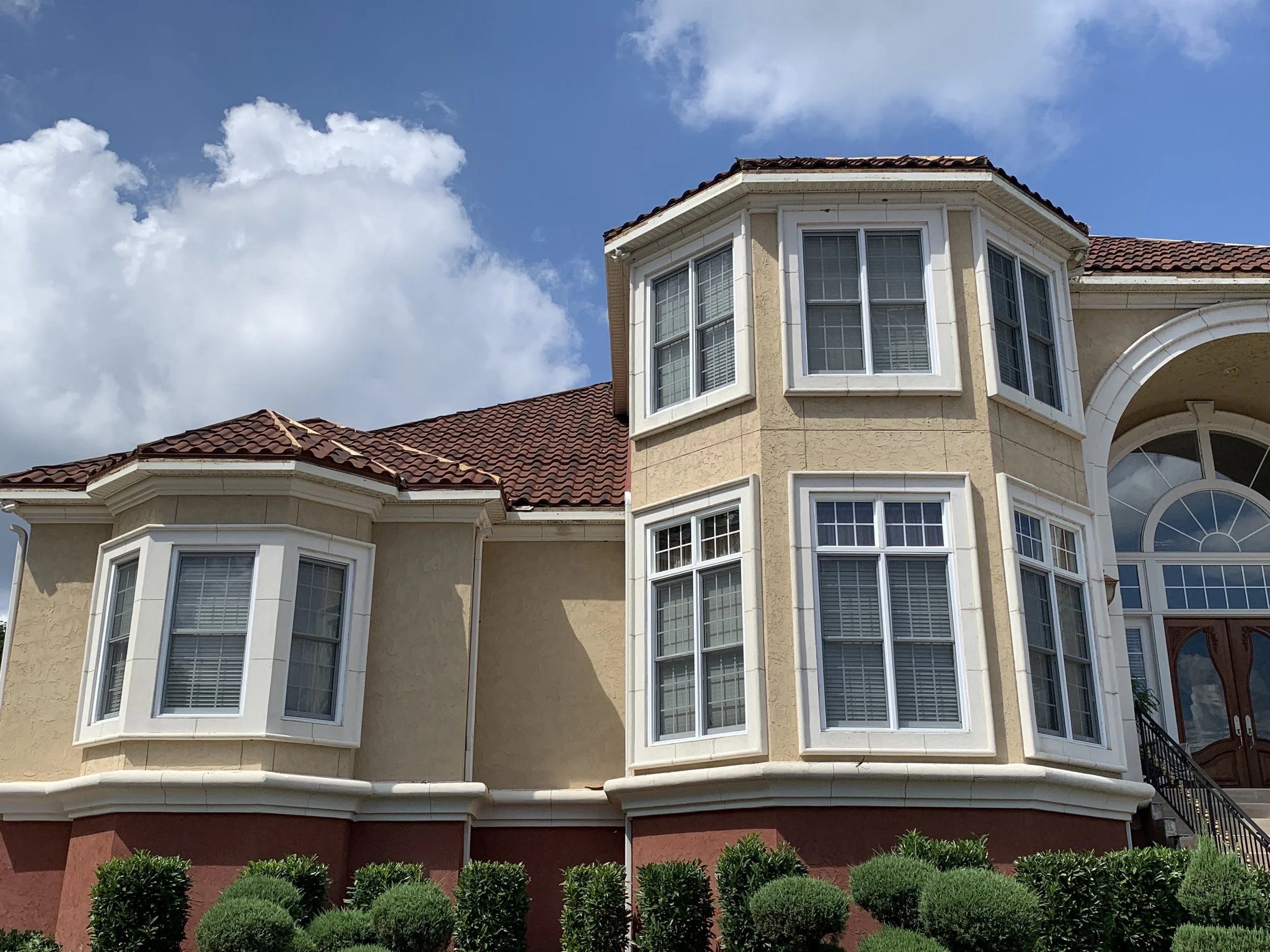 Close-up view of a large, multi-story house with beige stucco walls, white window trim, and a red-tiled roof, with a bright blue sky and scattered clouds in the background.