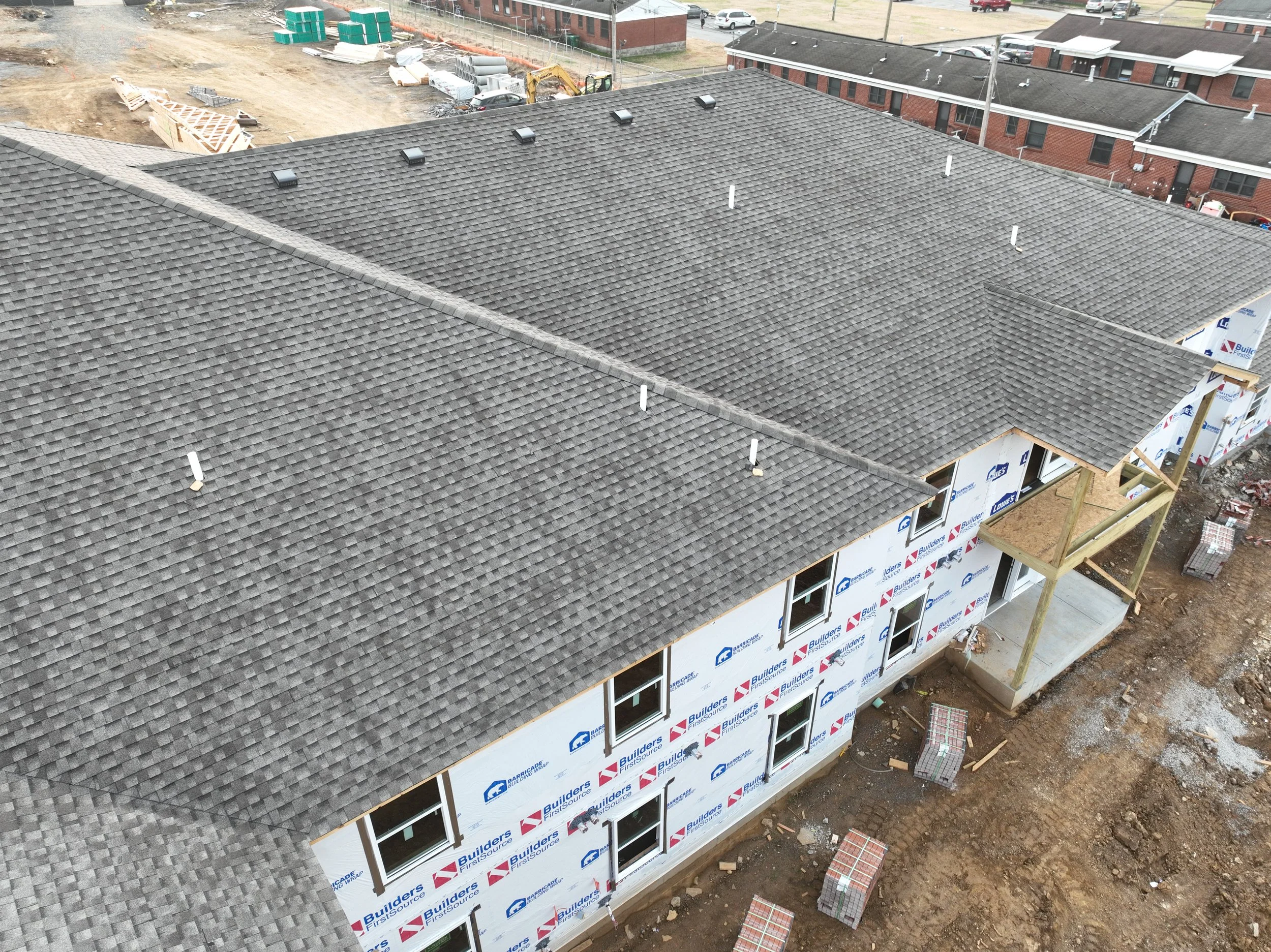 Aerial view of a house under construction with a completed dark gray shingle roof, surrounded by construction materials and equipment, with neighboring completed red brick homes.