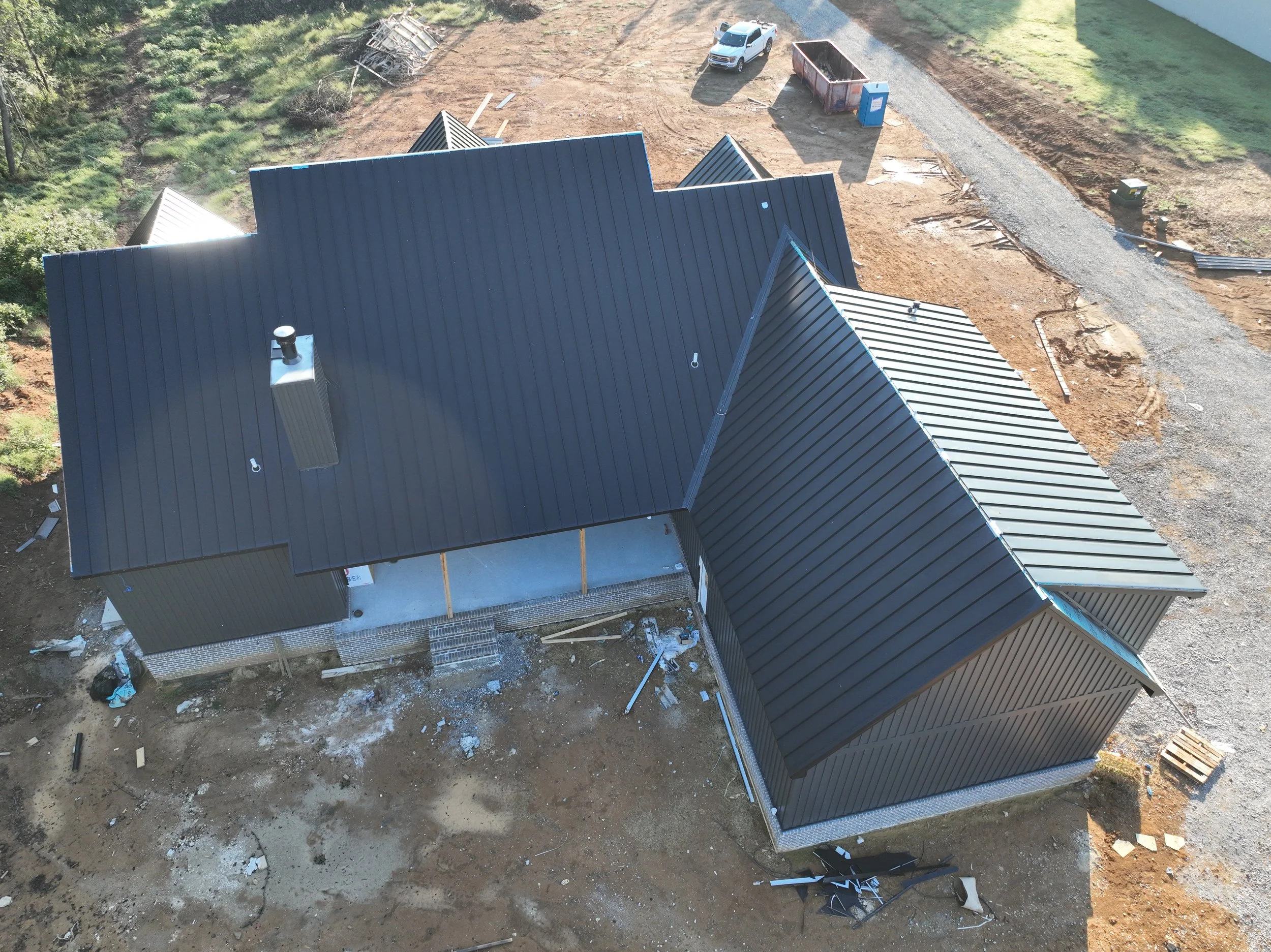Aerial view of a house under construction with a black metal roof, construction debris on the ground, a gravel driveway, a white pickup truck, and a blue portable toilet nearby.