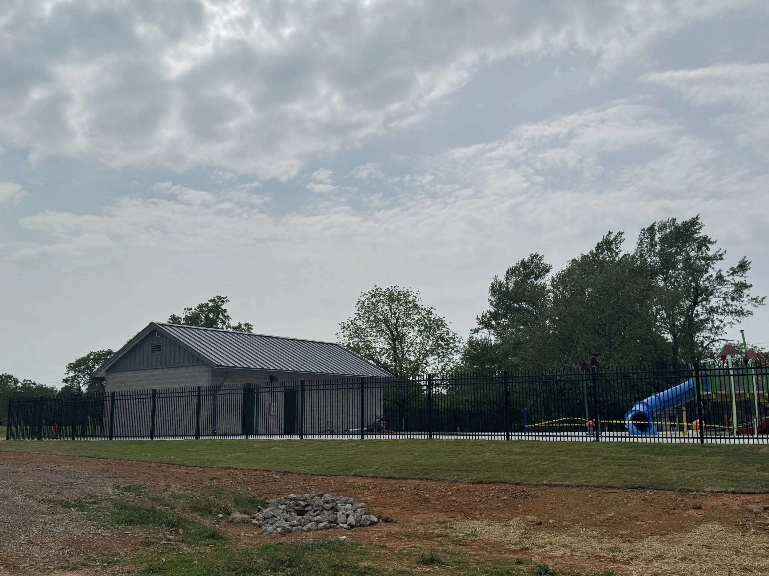 A backyard with a fenced play area, a house with a metallic roof, and trees. The sky is cloudy.