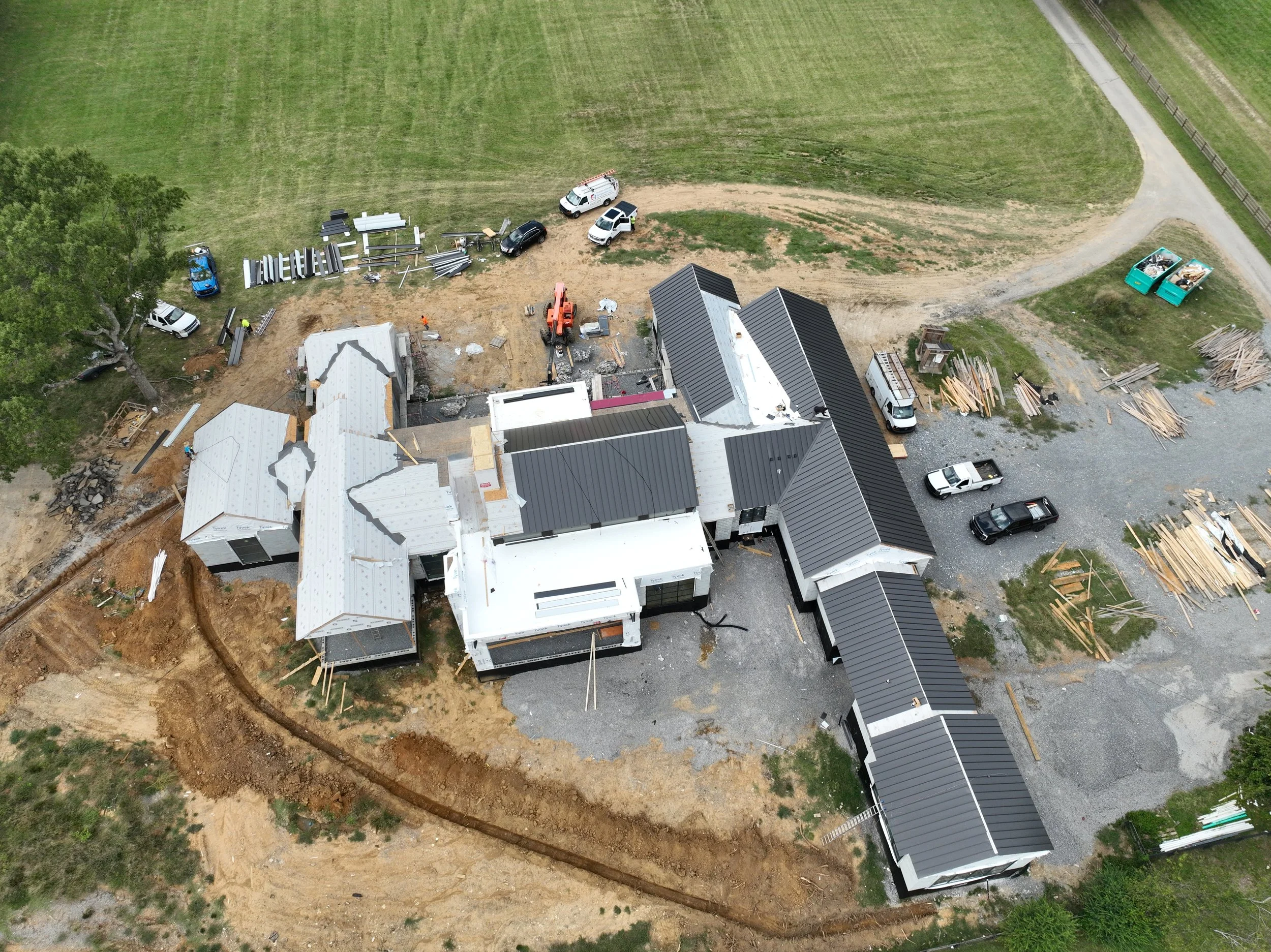 An aerial view of a construction site next to a grassy field. The site has building materials, construction vehicles, and partially constructed houses with metal roofs.