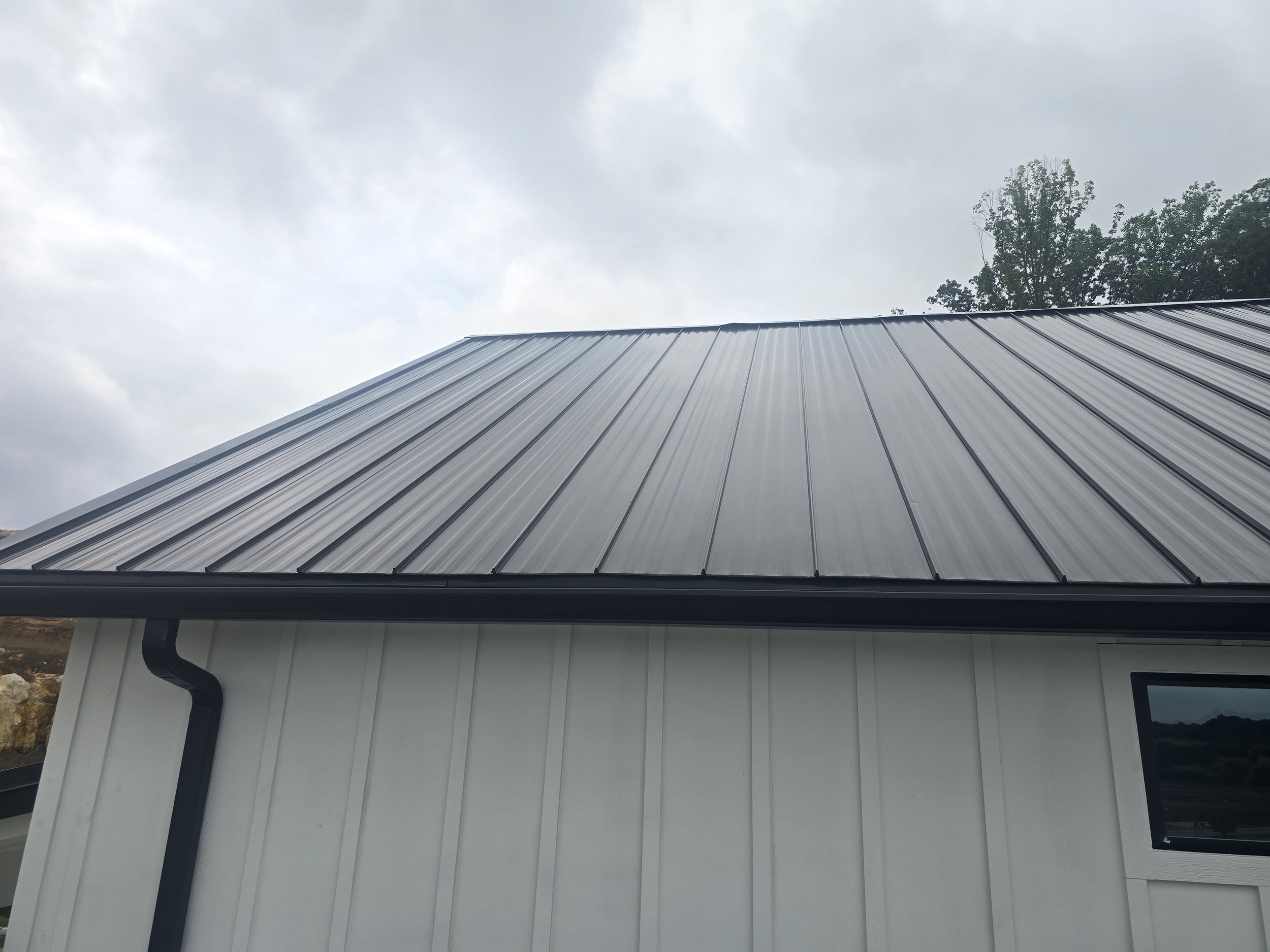 A building with white corrugated metal siding and a black gutter system, seen from a low angle against a cloudy sky with trees in the background.