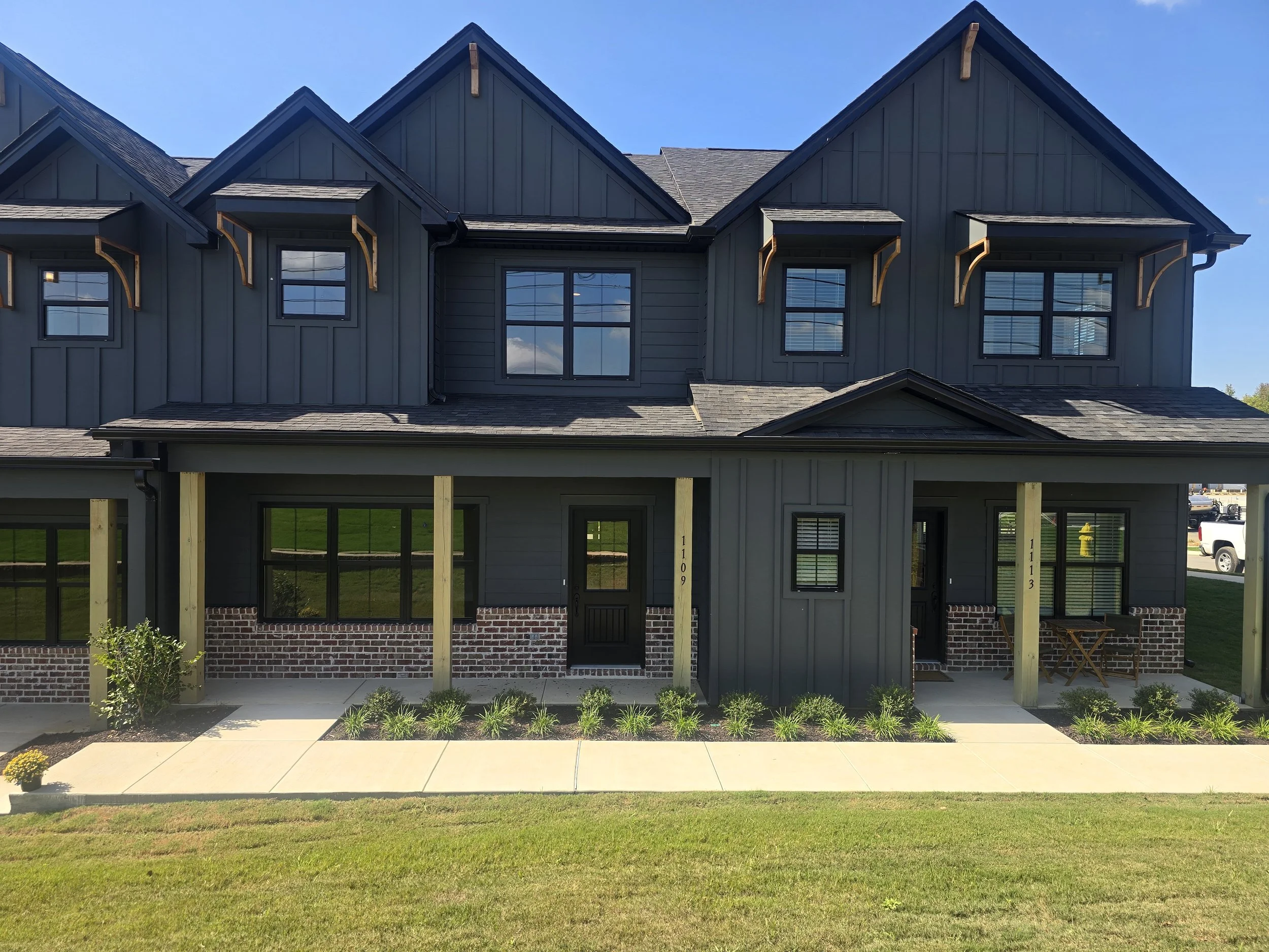 Front view of a modern, two-story house with gray siding and brick foundation. The house has multiple gabled roofs with wooden brackets under the eaves, and large windows. The front porch is under construction, supported by wooden posts, with a lands