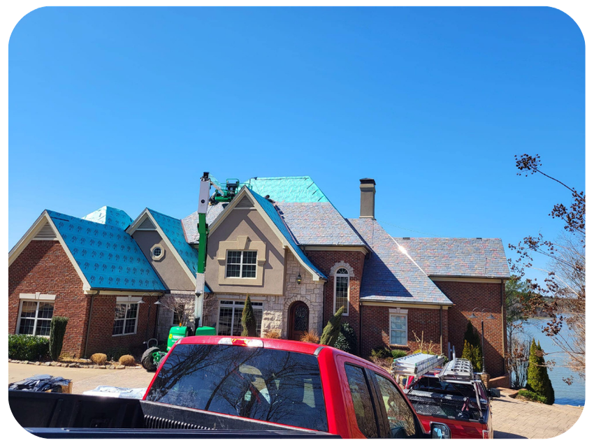 A house during roof construction with a crane lifting roofing materials. The house is brick with a steep roof, some parts are covered with blue and green roofing materials. A red truck is parked in front, and construction equipment is visible.