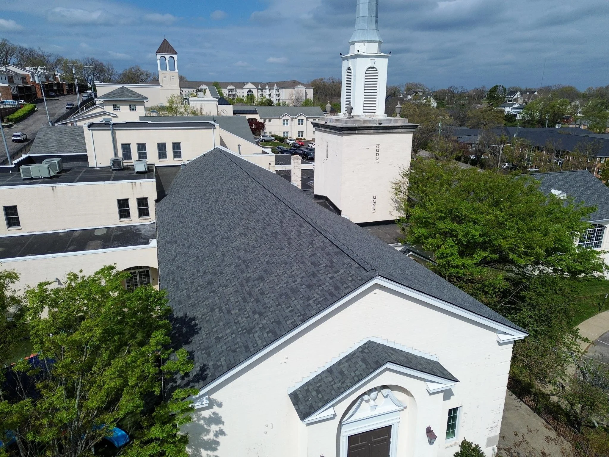 Bird's eye view of a large white church with a tall steeple, surrounded by residential buildings, trees, and parking lots on a partly cloudy day.