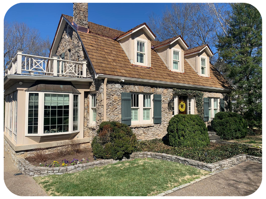 A charming house with stone and beige siding, green shutters, and a steep roof with dormer windows, surrounded by a landscaped yard with bushes and flowers.