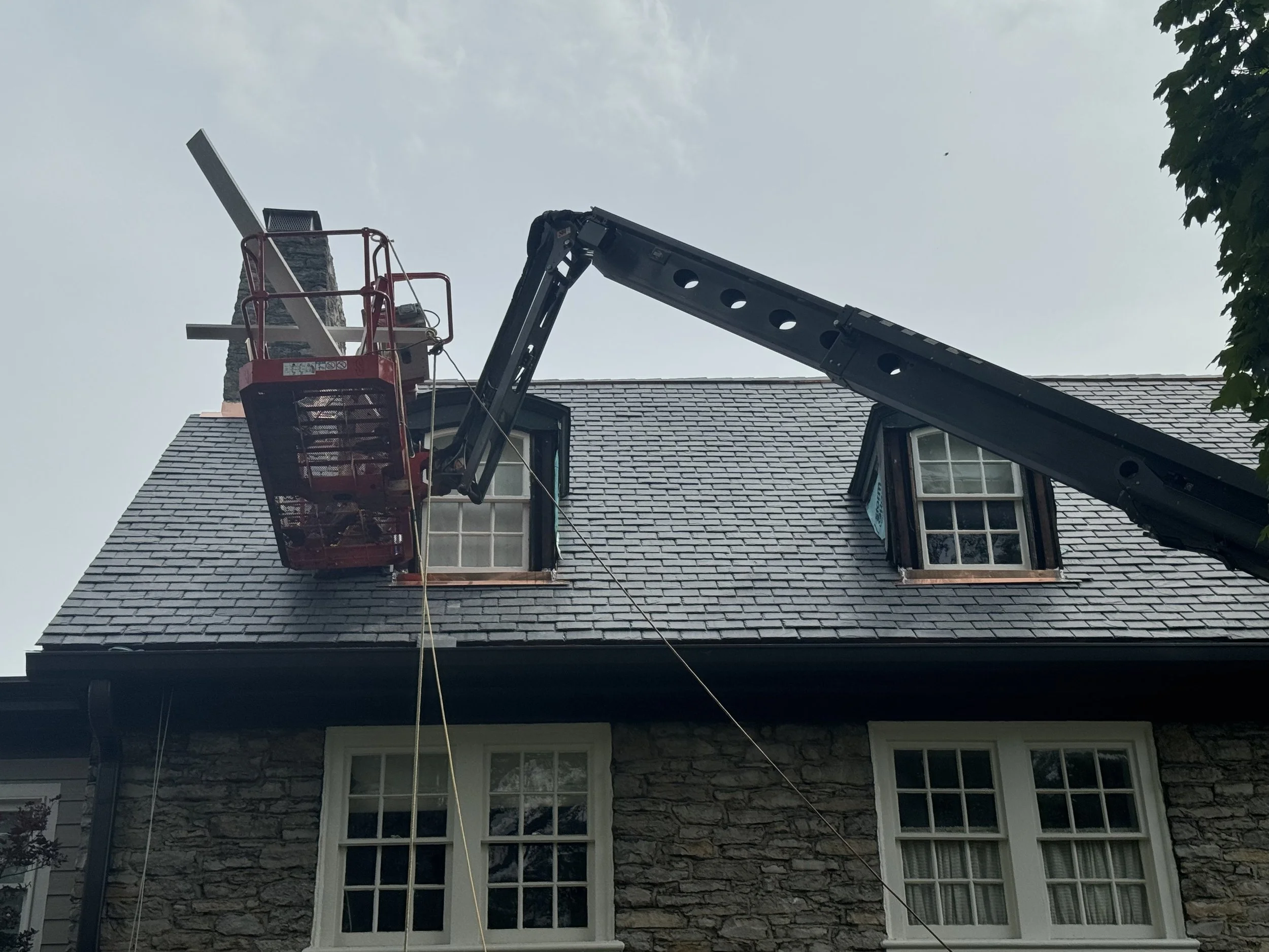 A house with gray shingle roof and large windows, with a ladder lift reaching the chimney. An operator's basket is attached to the boom of the lift, positioned near the chimney. Trees are visible on the right, and the sky is overcast with some clouds