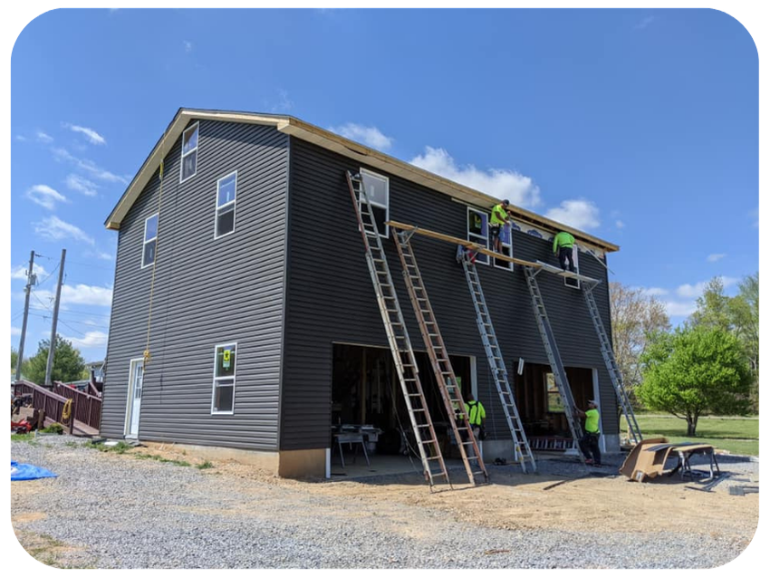 Construction workers installing a roof on a multi-story house, with ladders leaning against the building, on a clear day with blue sky and some trees in the background.