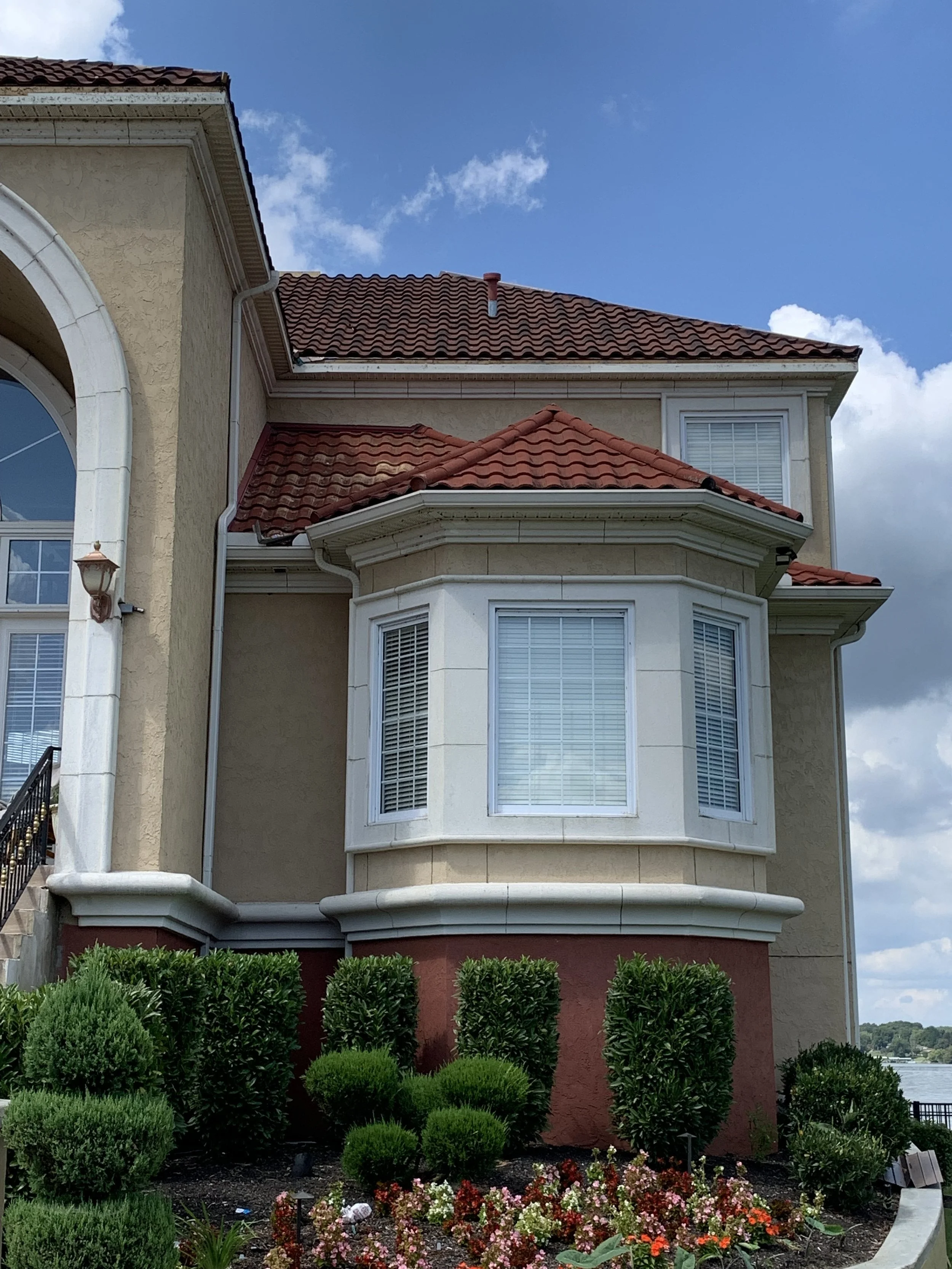 A multi-story house with beige stucco walls, red tiled roof, and a bay window at the front. There is a garden with neatly trimmed bushes and colorful flowers in front of the house, and the sky is partly cloudy.