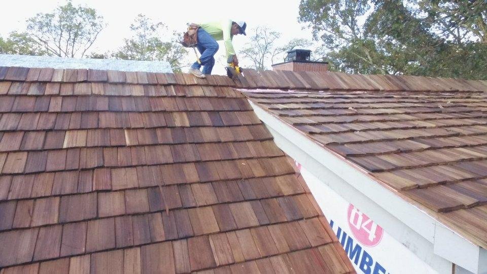 A worker installing or repairing roof shingles on a house, standing on the roof and using tools, with trees visible in the background.