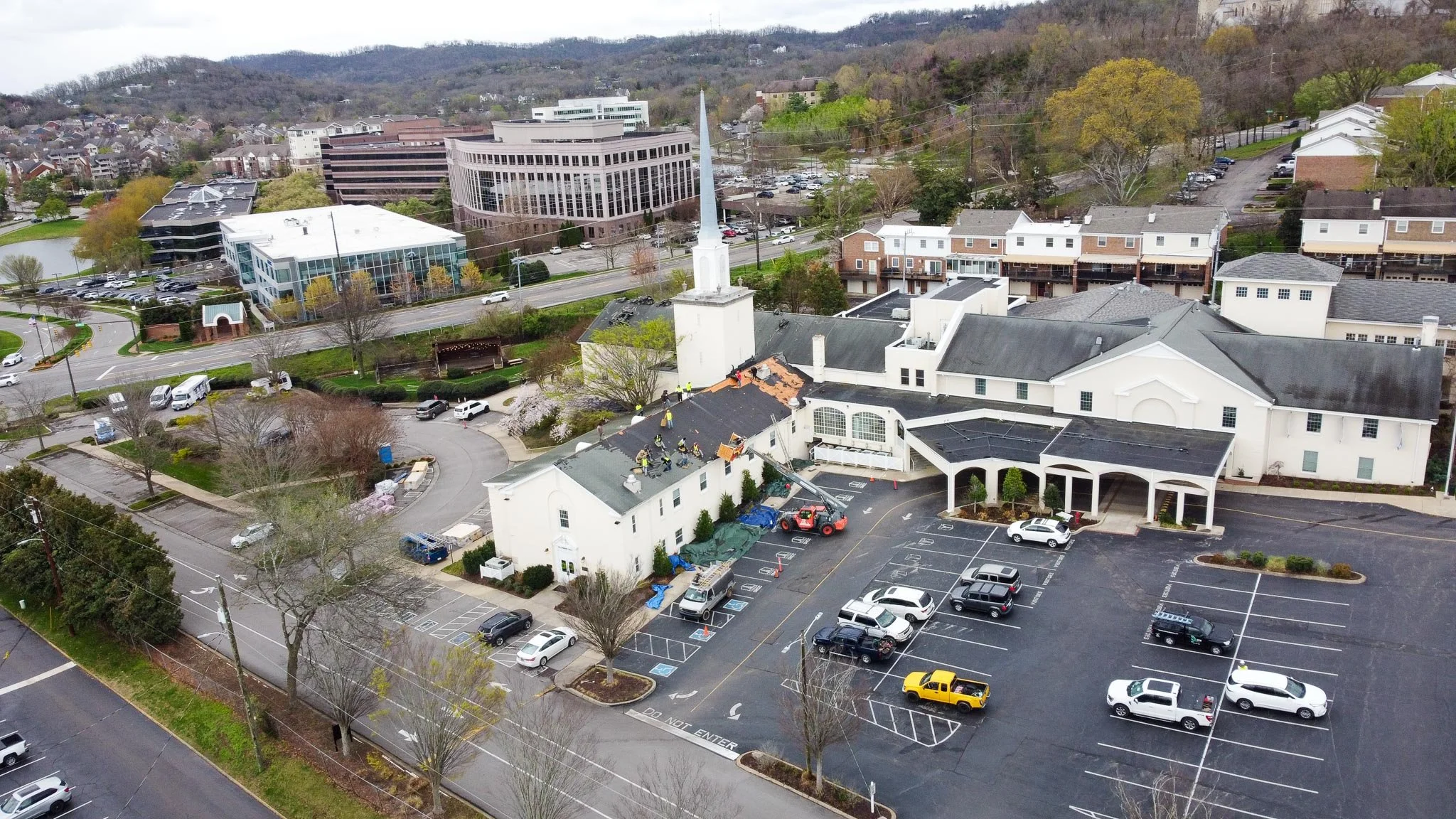 An aerial view of a church with workers on the roof repairing damage, surrounded by parking lot with cars, nearby office buildings, and residential houses against a hilly landscape.